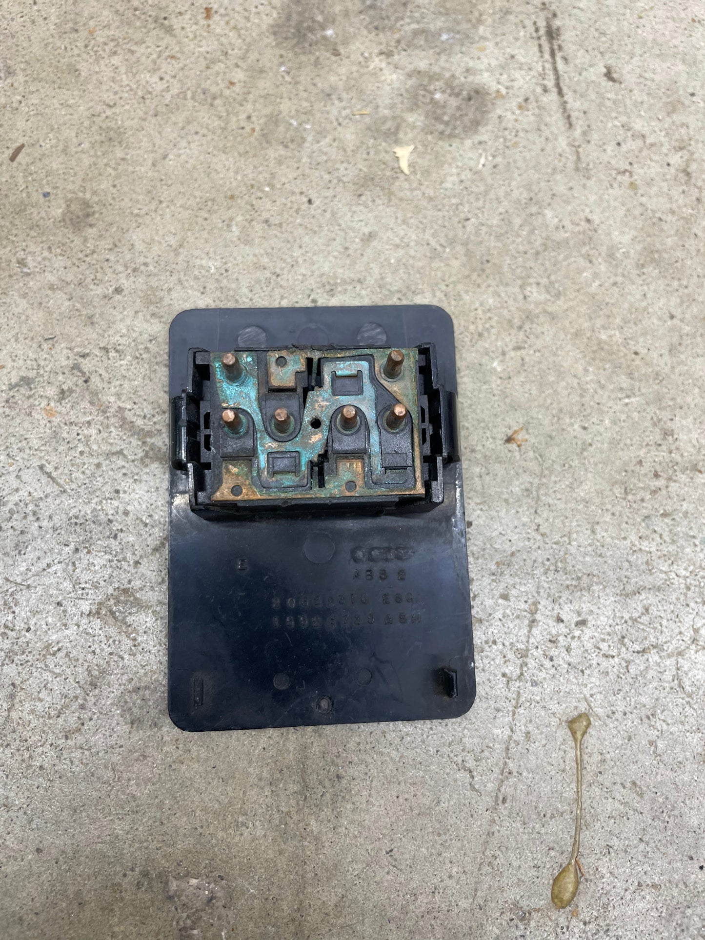 A close-up of a General Motors OEM Driver Side Window Switch for 1986-94 Chevy S10, Blazer, and Syclone shows corroded copper connectors on its black plastic base, placed on rough gray concrete.