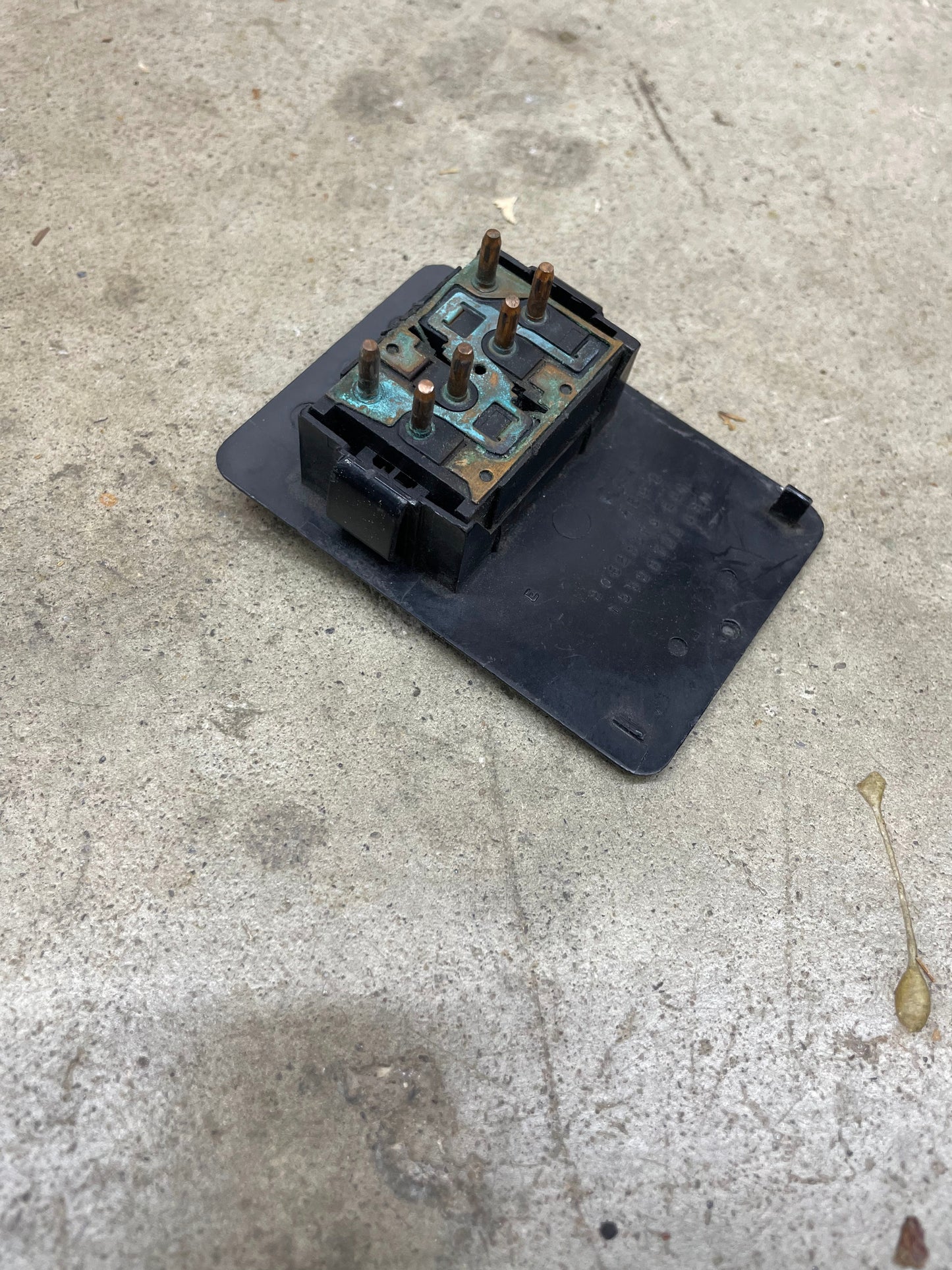 A rectangular black General Motors OEM driver side window switch for 1986-94 Chevy S10, Blazer, Syclone, and more shows corroded metal prongs as it rests on a rough, stained concrete surface.