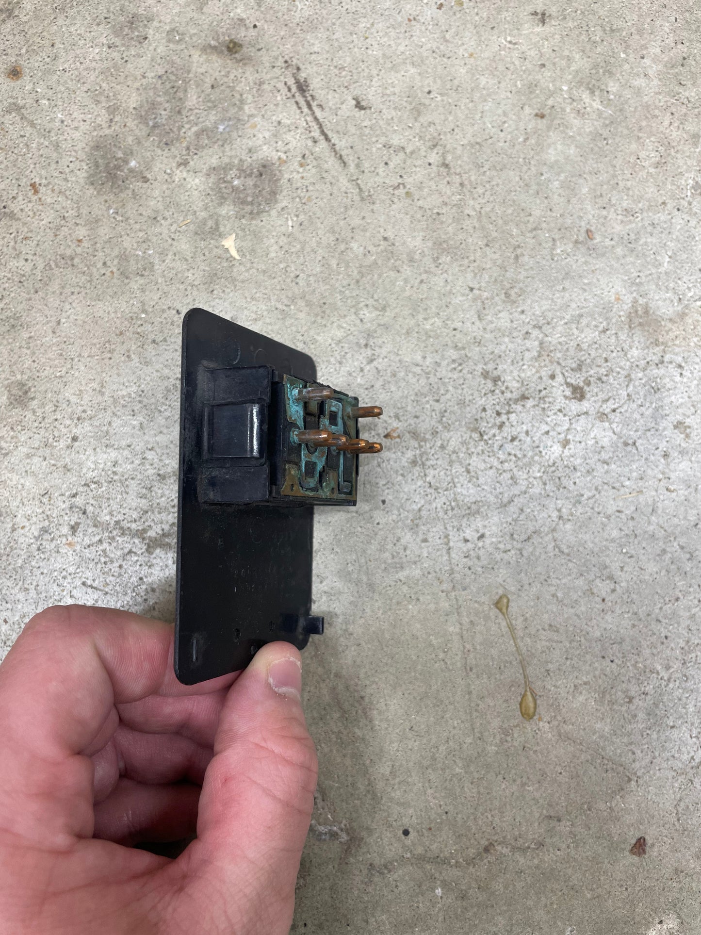 A hand holds an OEM Driver Side Window Switch for 2 Door 1986-94 Chevy S10, Blazer, Syclone, and more by General Motors, featuring a black plastic base against a concrete surface background.
