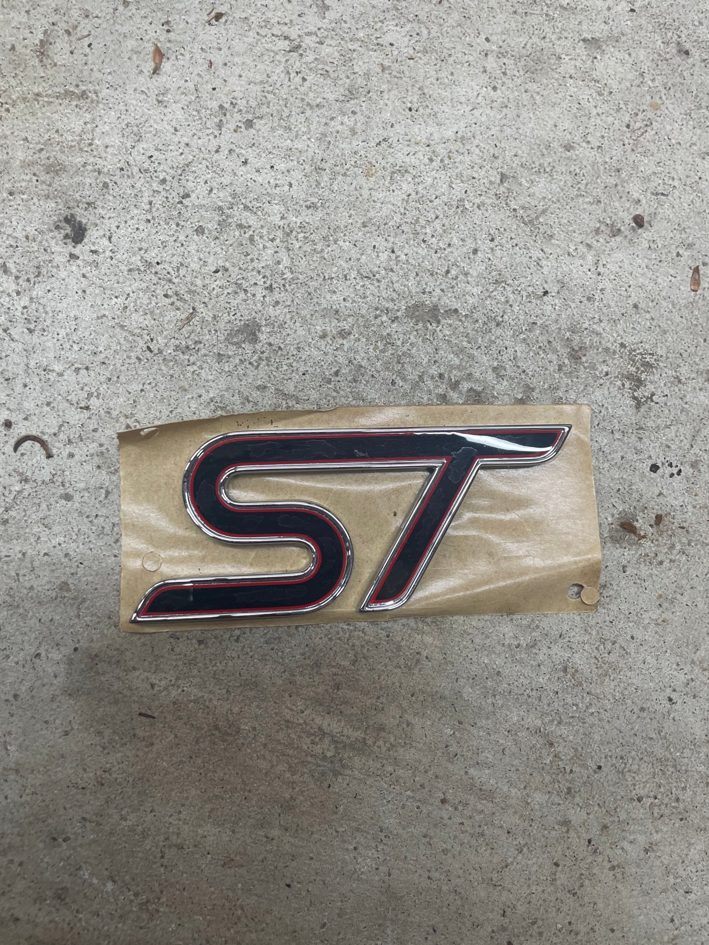 A NOS Rare Limited Ford ST Octane Academy Badge Emblem Set for 2012-2018 Focus/Fiesta by General Motors rests on brown paper atop rough concrete.