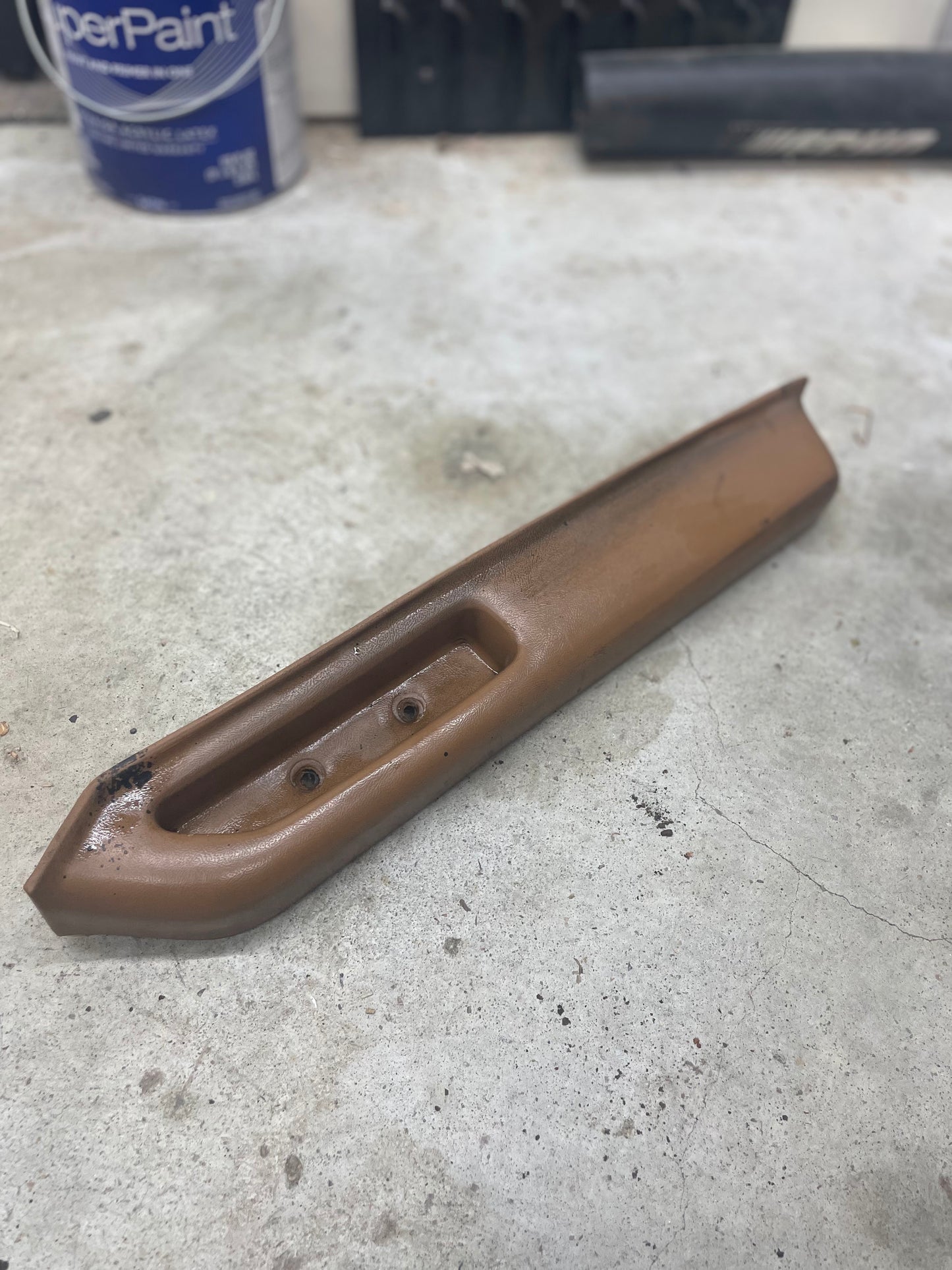 A beige OEM passenger door panel armrest handle by General Motors for 1986-1993 Chevy S10, Blazer, and more, with screw holes, rests on a concrete floor. A blue Super Paint can and other workshop items are visible in the background.