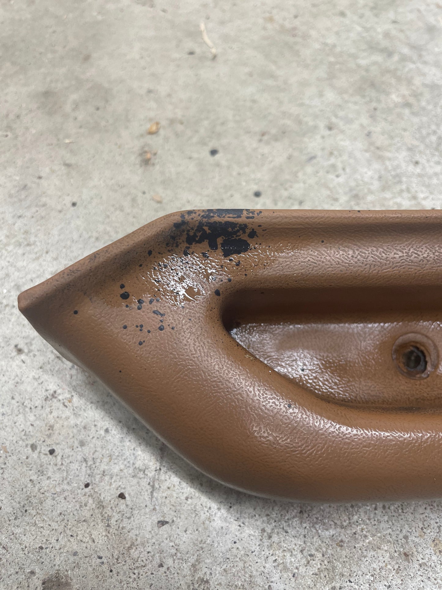 Close-up of a beige OEM passenger door panel armrest handle by General Motors for 1986-1993 Chevy S10, Blazer, and more, showing black paint splatters on one end, resting on rough gray concrete.