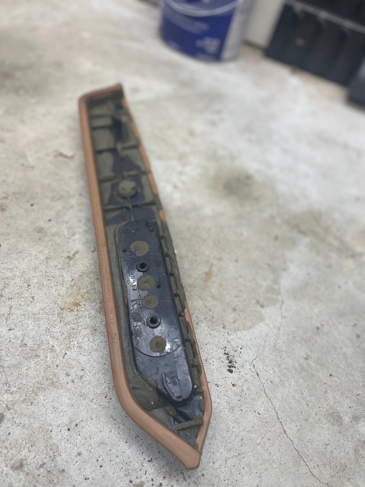 A used OEM beige passenger door panel armrest handle by General Motors for 1986-1993 Chevy S10, Blazer, and more lies on a stained concrete floor in a workshop, showing grime and visible mounting holes.