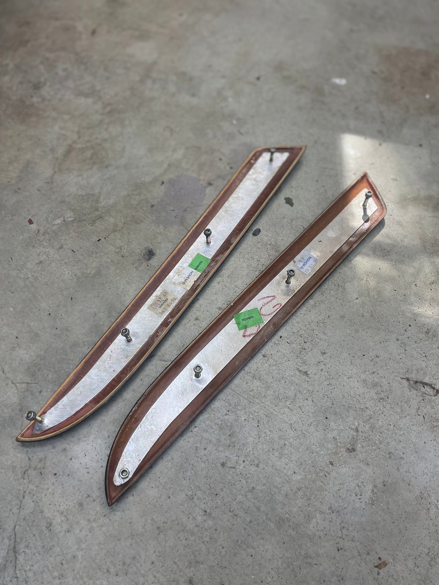A pair of OEM General Motors front door panel woodgrain trim inserts with cracks for 2002-2006 Cadillac Escalade lies on a concrete floor, showing attached metal bolts, green labels, and visible shadows and light spots in the background.