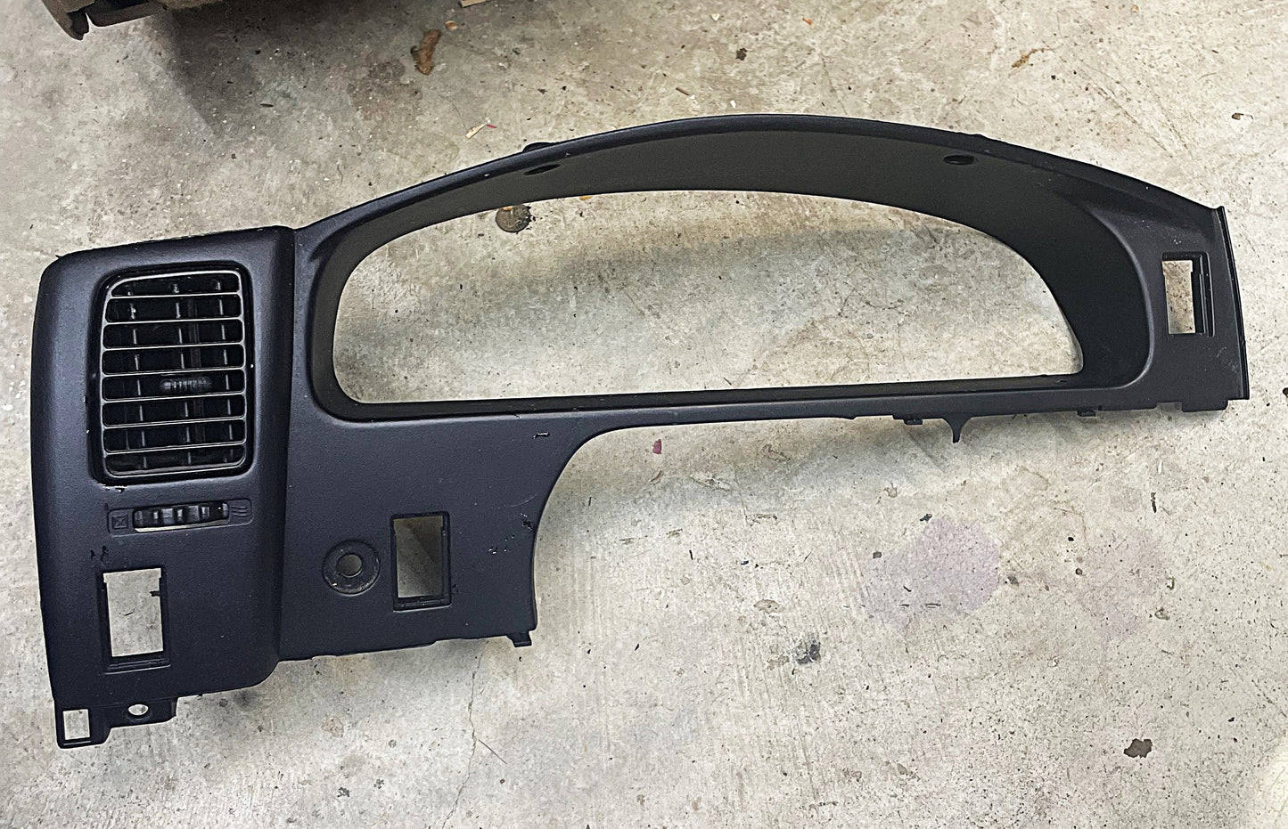 A General Motors OEM Instrument Gauge Cluster Dash Bezel for 1995-2004 Toyota Tacoma, featuring vent, button slots, and cutouts, is shown lying on a concrete floor.