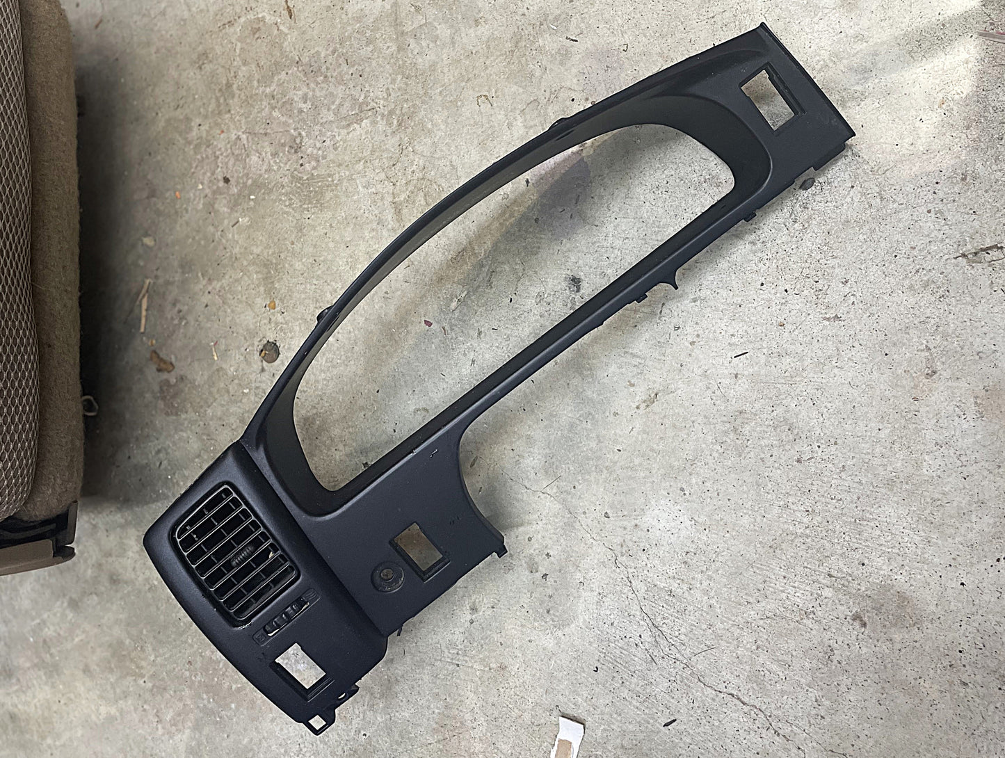 A General Motors OEM Instrument Gauge Cluster Dash Bezel for 1995-2004 Toyota Tacoma is lying on a concrete floor next to a brown fabric seat.