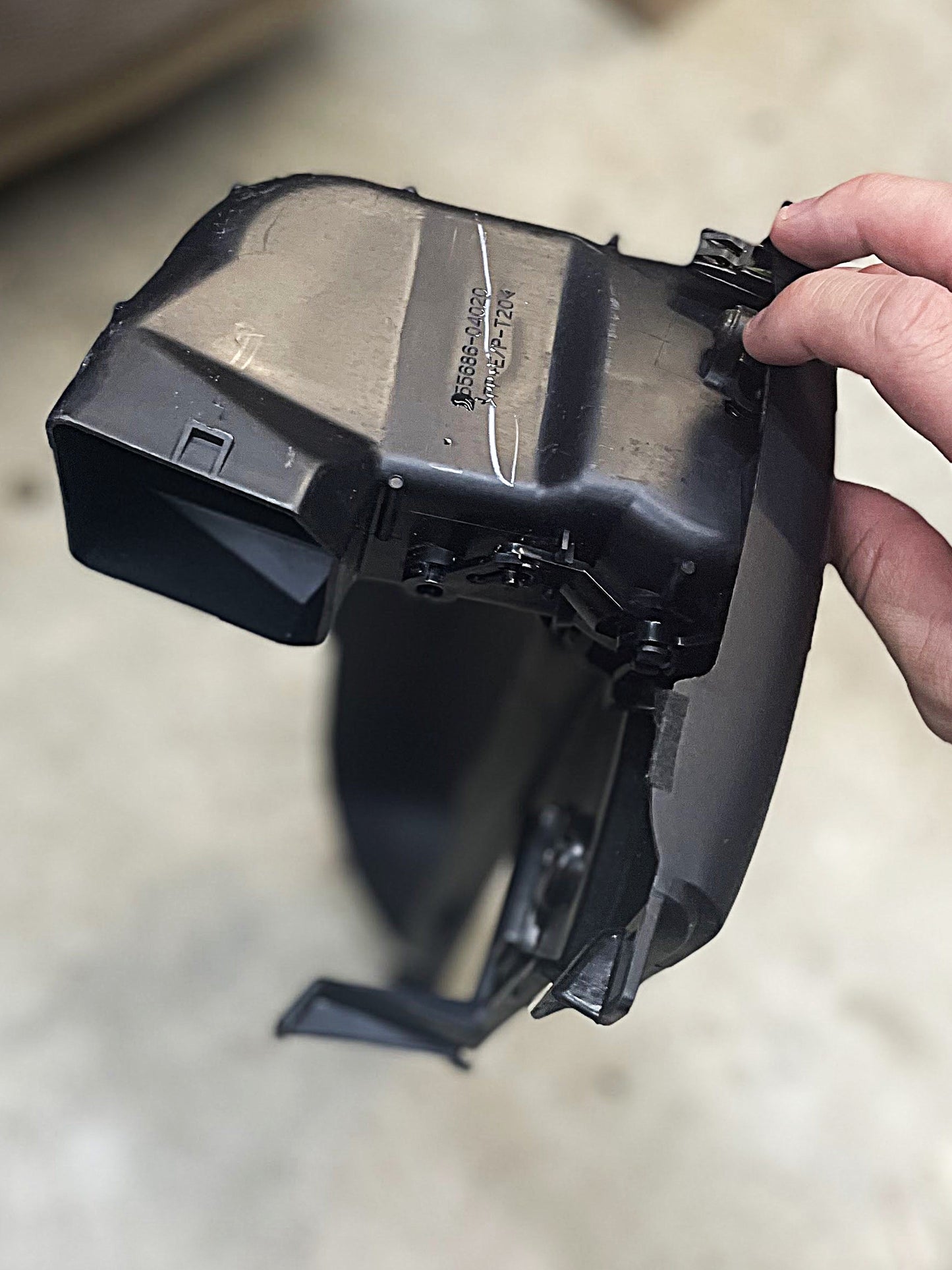 A person holds a General Motors OEM Instrument Gauge Cluster Dash Bezel for 1995-2004 Toyota Tacoma, with a scratched black surface and visible clips, set against a blurred indoor background.