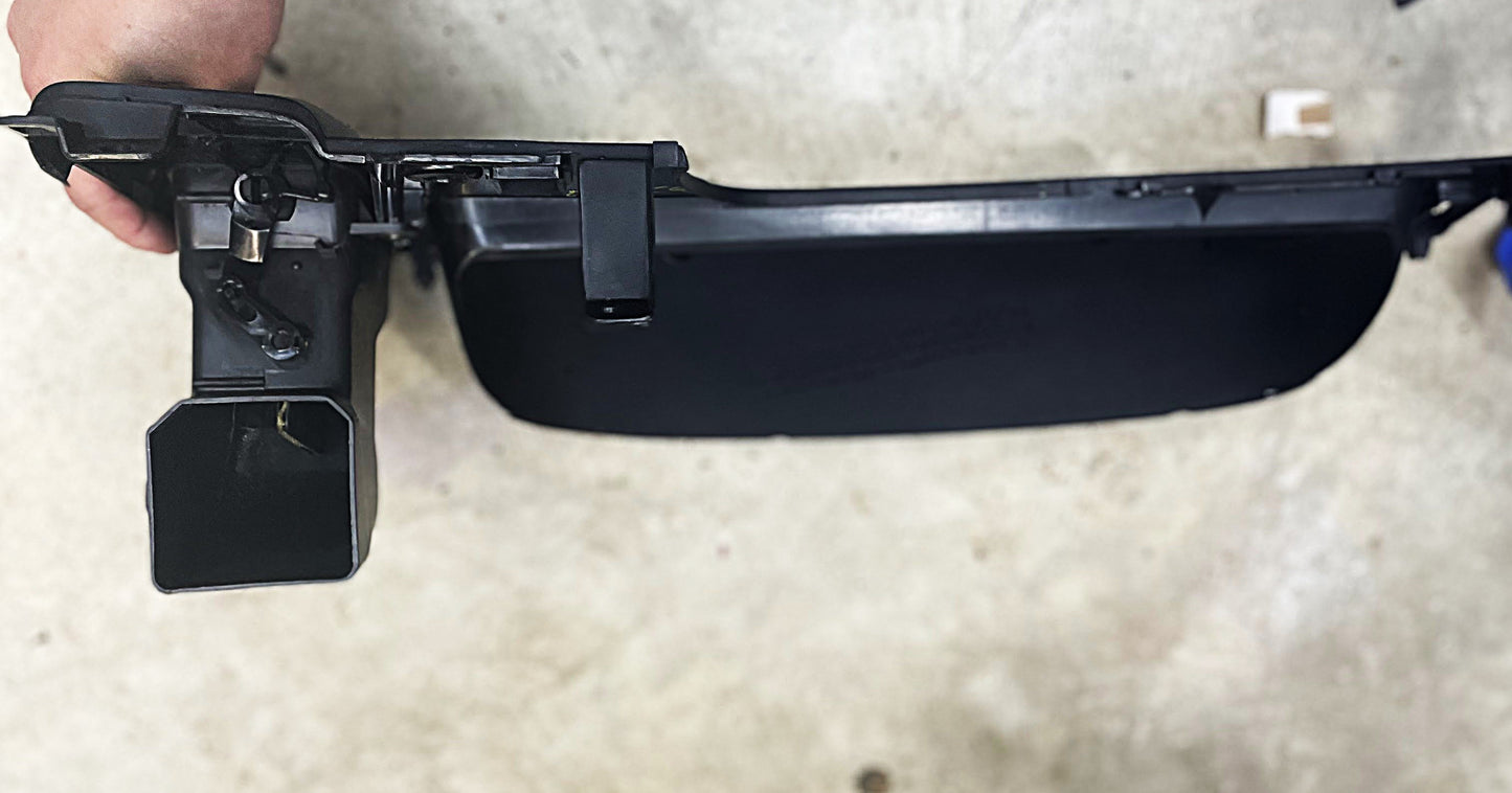 A person holds a General Motors OEM Instrument Gauge Cluster Dash Bezel for 1995-2004 Toyota Tacoma against a light, textured surface. This black rectangular bezel features multiple sections and attached components.