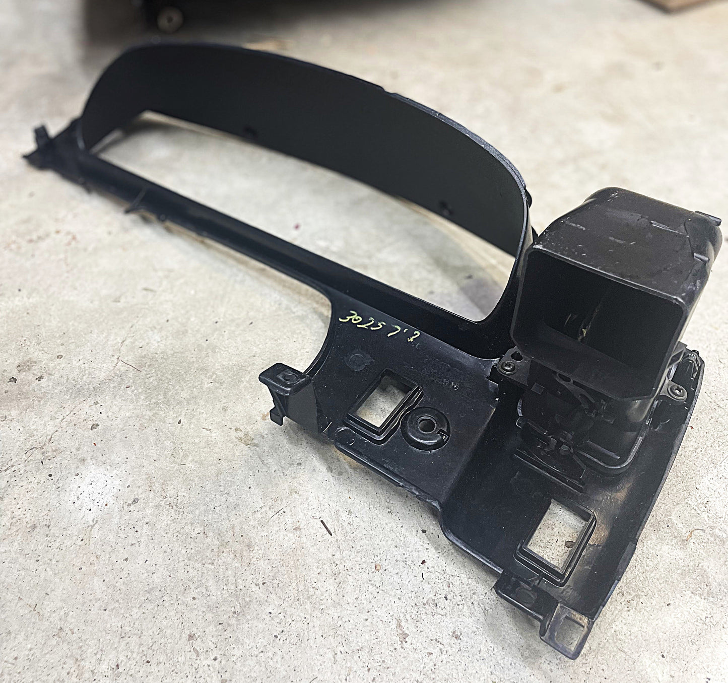 A black plastic OEM Instrument Gauge Cluster Dash Bezel by General Motors for 1995-2004 Toyota Tacoma, with yellow handwritten markings and cutouts, rests on a concrete floor.