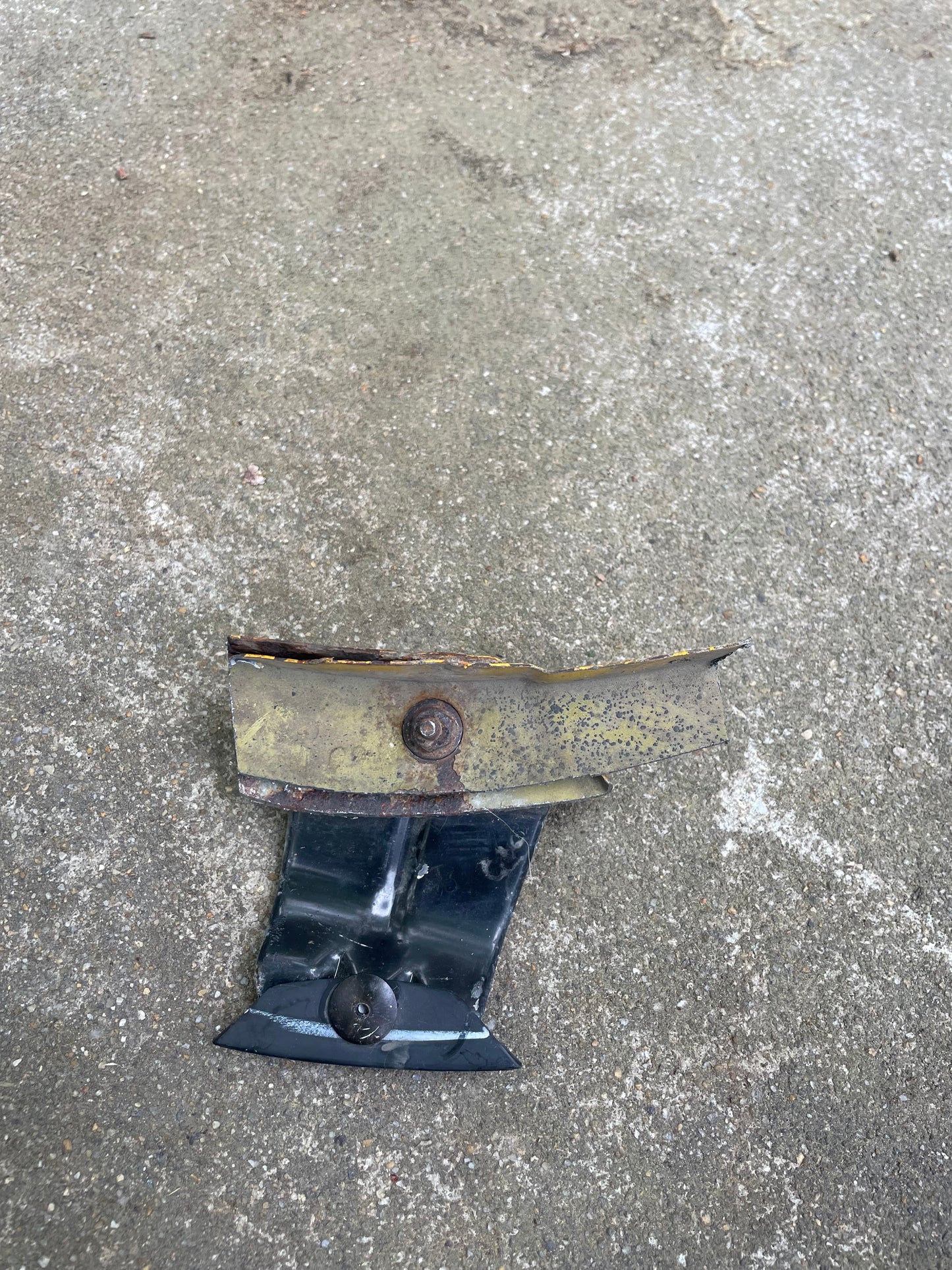 A small, rusted metal object with bolts and chipped paint rests on rough gray concrete. It appears to be part of a General Motors OEM Rear Left Driver ZR2 Fender Flare Body Side Extension for 1995-2005 Chevy Blazer or GMC Jimmy.