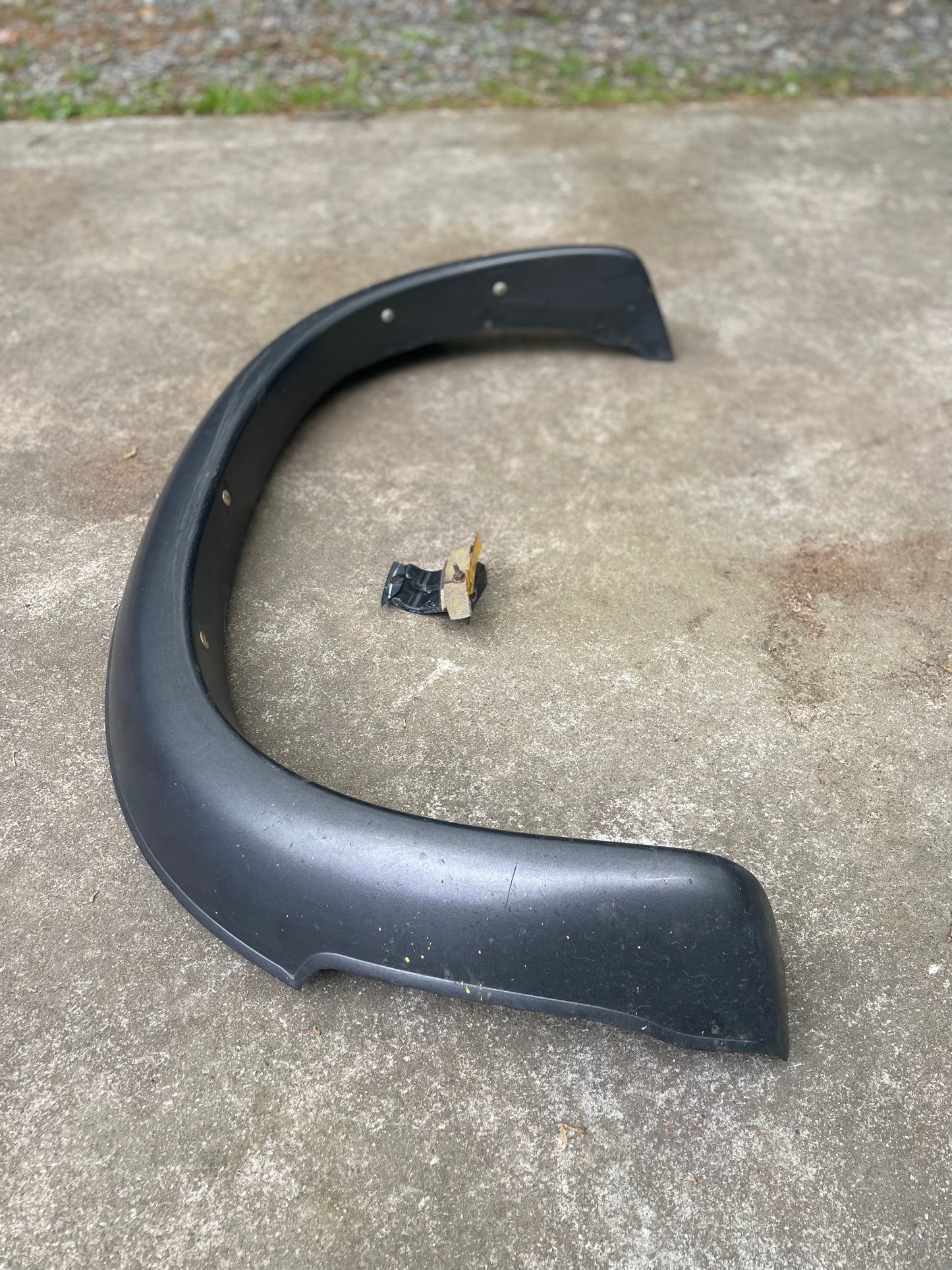 A General Motors OEM Rear Left Driver ZR2 Fender Flare Body Side Extension for 1995-2005 Chevy Blazer or GMC Jimmy is shown on concrete with a small black and beige mounting bracket inside its arc; grass and gravel appear in the background.