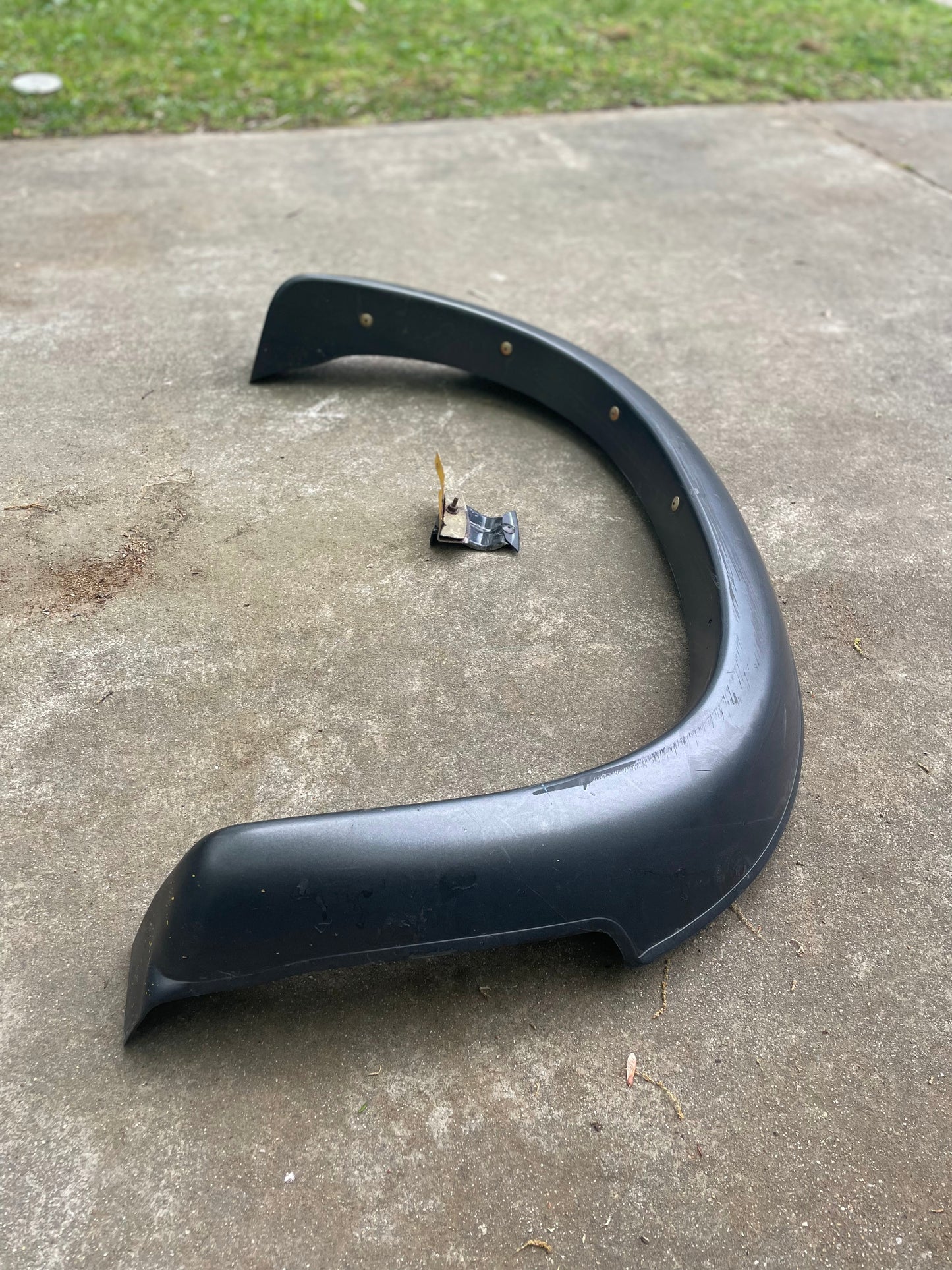 A General Motors OEM Rear Left Driver ZR2 Fender Flare Body Side Extension for 1995-2005 Chevy Blazer or GMC Jimmy rests on a concrete surface outdoors, with included hardware in its curve and grass visible in the background.