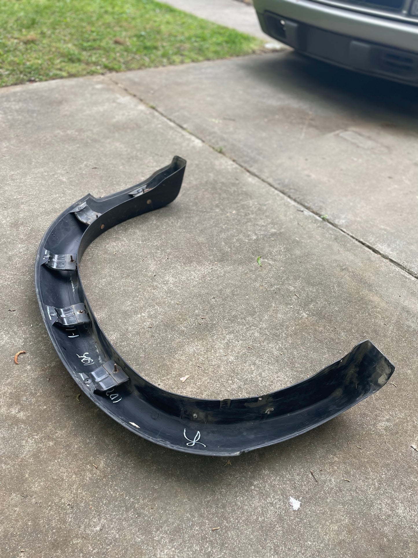 A General Motors OEM Rear Left Driver ZR2 Fender Flare Body Side Extension for 1995-2005 Chevy Blazer/GMC Jimmy rests on a concrete driveway, with grass and a parked vehicle in the background.