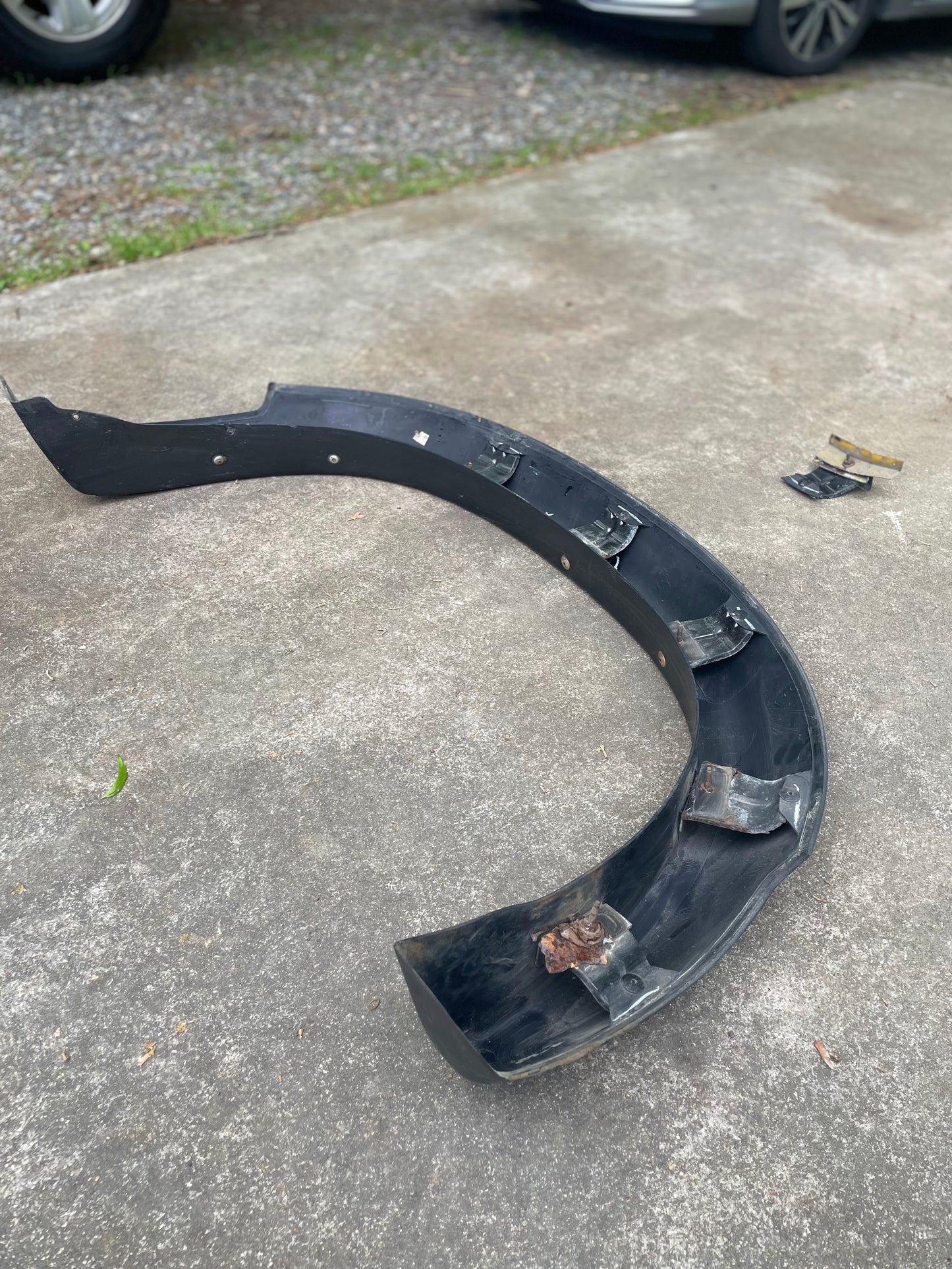 An OEM Rear Left Driver ZR2 Fender Flare Body Side Extension by General Motors for 1995-2005 Chevy Blazer or GMC Jimmy sits on a concrete driveway with metal brackets attached and two detached brackets nearby. A car and gravel are visible in the background.