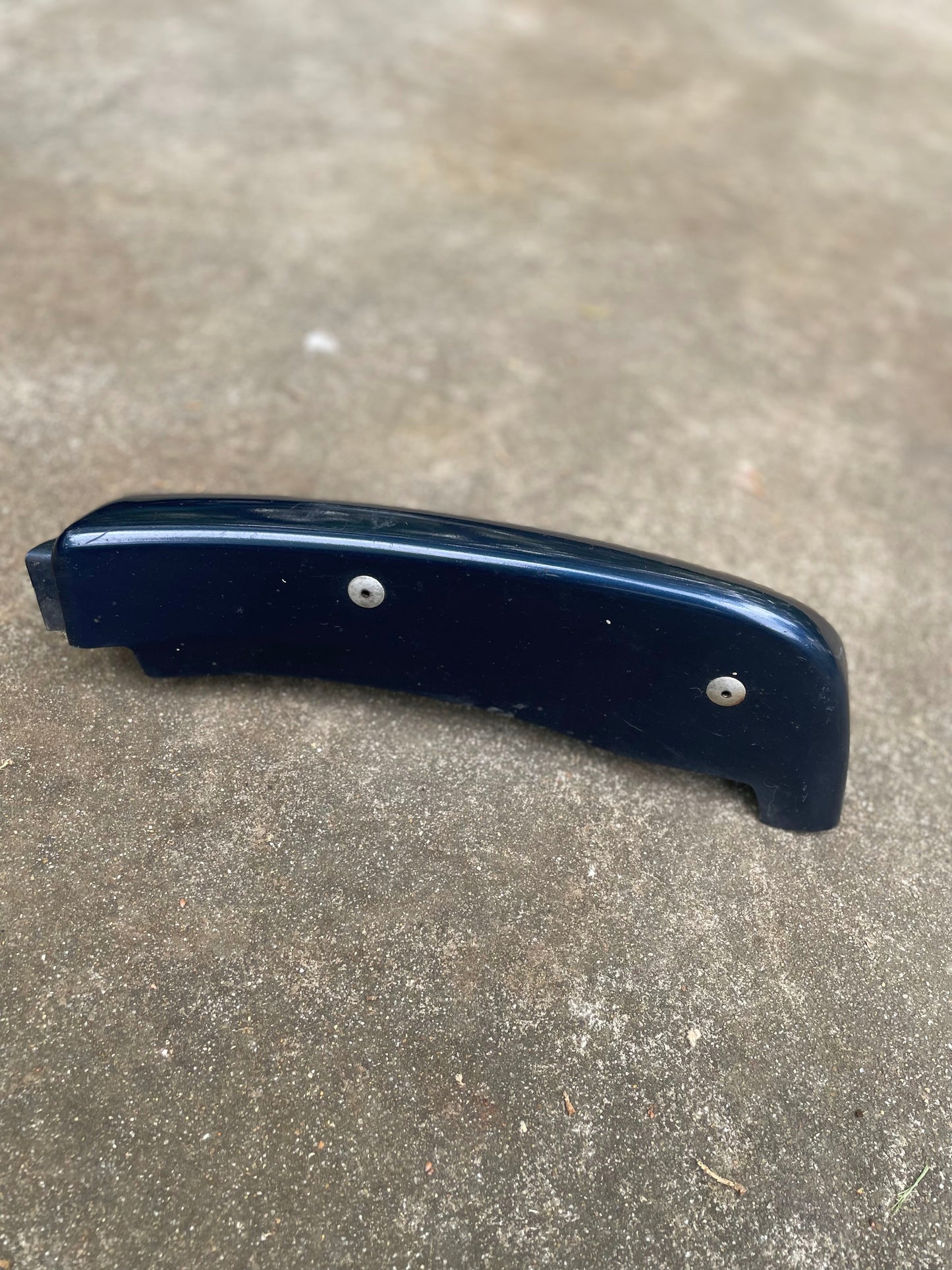 A General Motors OEM Front Right Passenger ZR2 Bumper Flare Extension in Blue for 1998-2005 GMC Sonoma and Jimmy is lying on a rough concrete surface. Two screws are visible beside the curved plastic part.