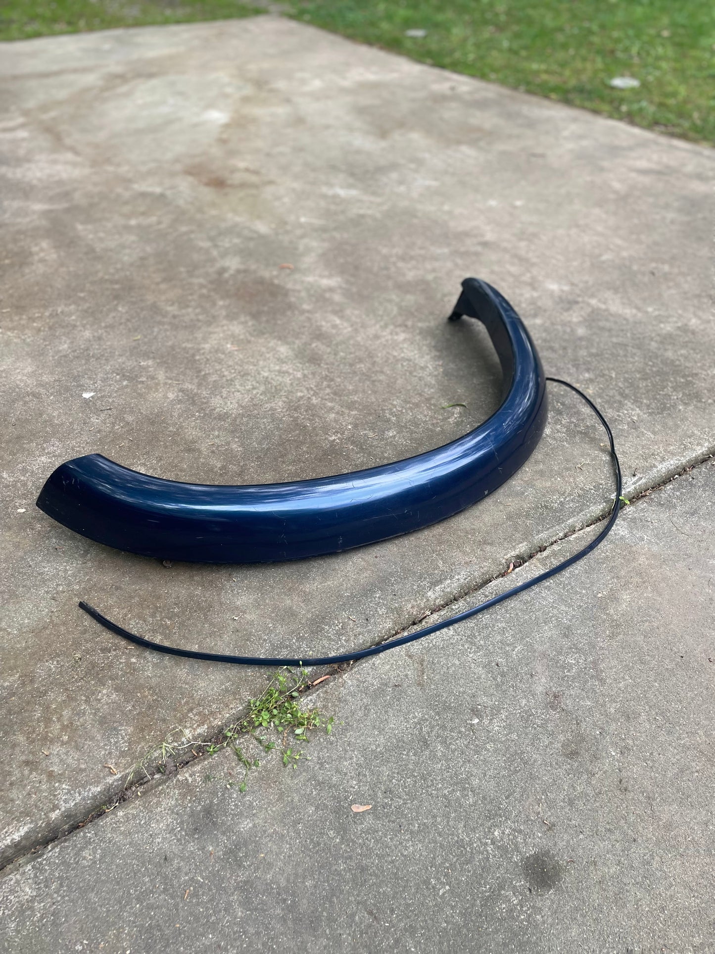 A General Motors OEM Front Right Passenger ZR2 Fender Flare Extension in blue for 1994-2005 Chevy S10/GMC Sonoma lies curved on a concrete driveway beside a thin black strip, with green grass in the background.