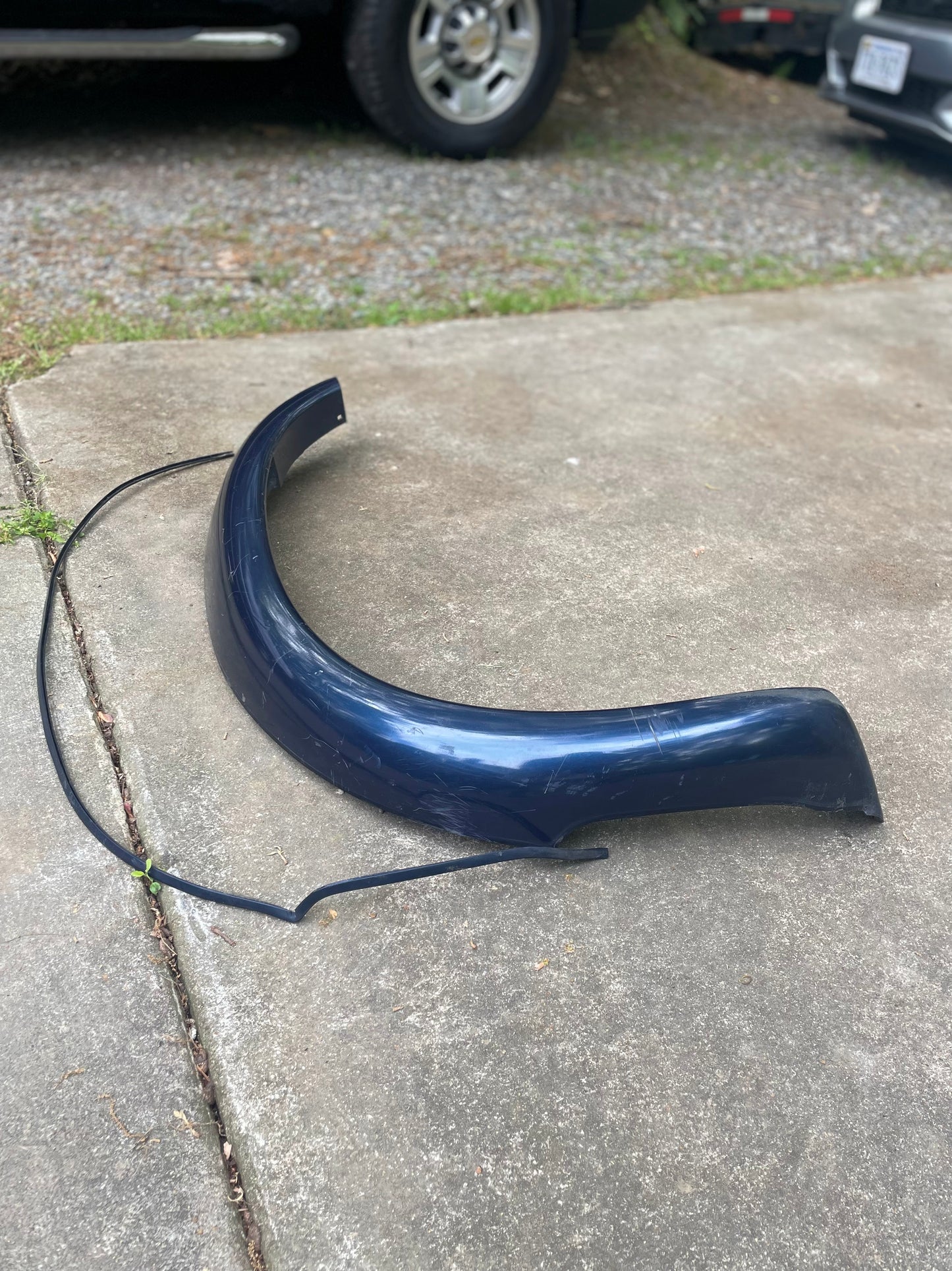 A blue OEM General Motors Front Right Passenger ZR2 Fender Flare Extension for 1994-2005 Chevy S10, GMC Sonoma, etc. lies scratched on a concrete driveway; other vehicles and gravel appear in the background.