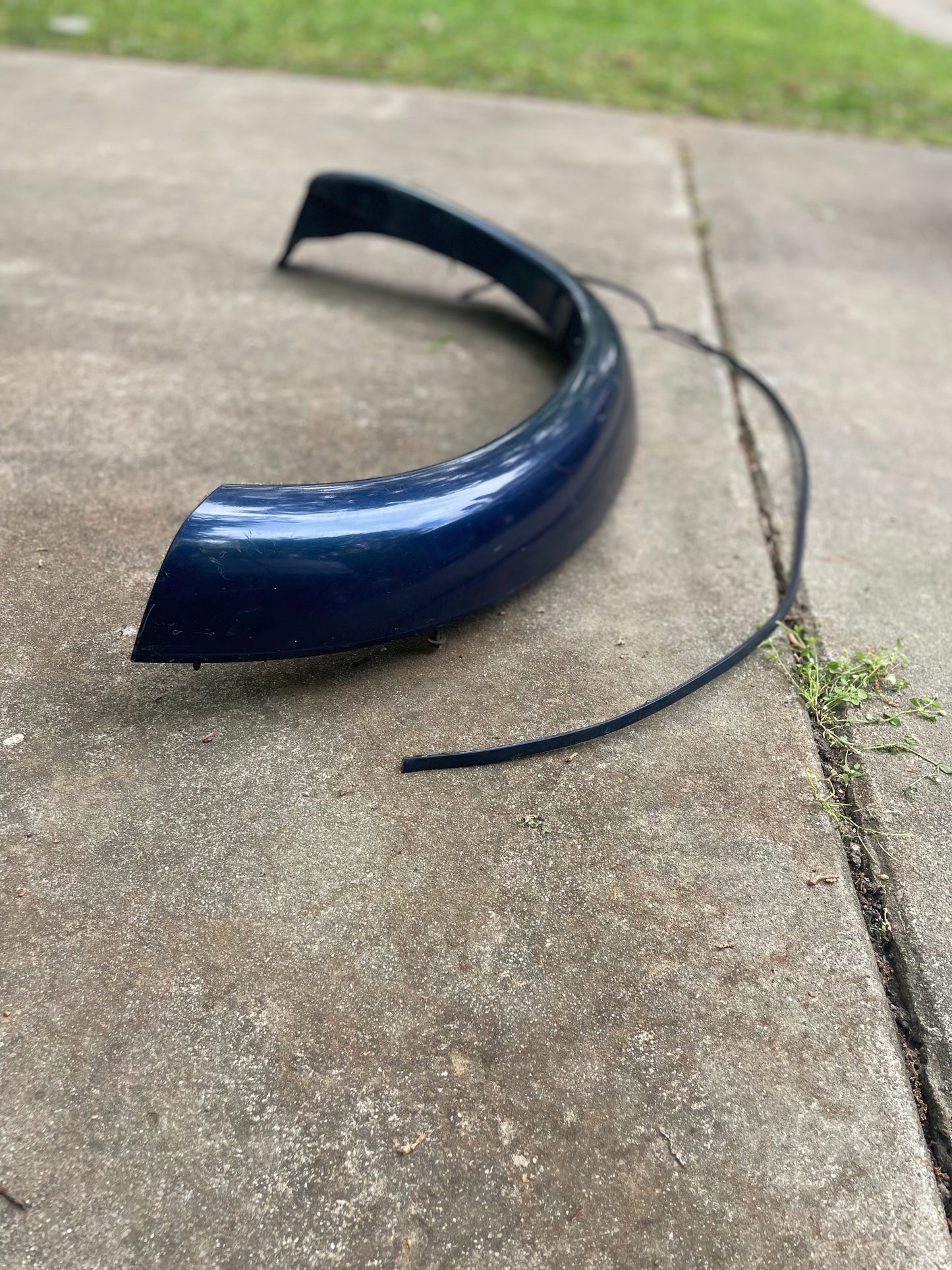 A General Motors OEM Front Right Passenger ZR2 Fender Flare Extension in blue for 1994-2005 Chevy S10 and GMC Sonoma rests on a concrete driveway, with a detached black trim piece nearby and grass in the background.