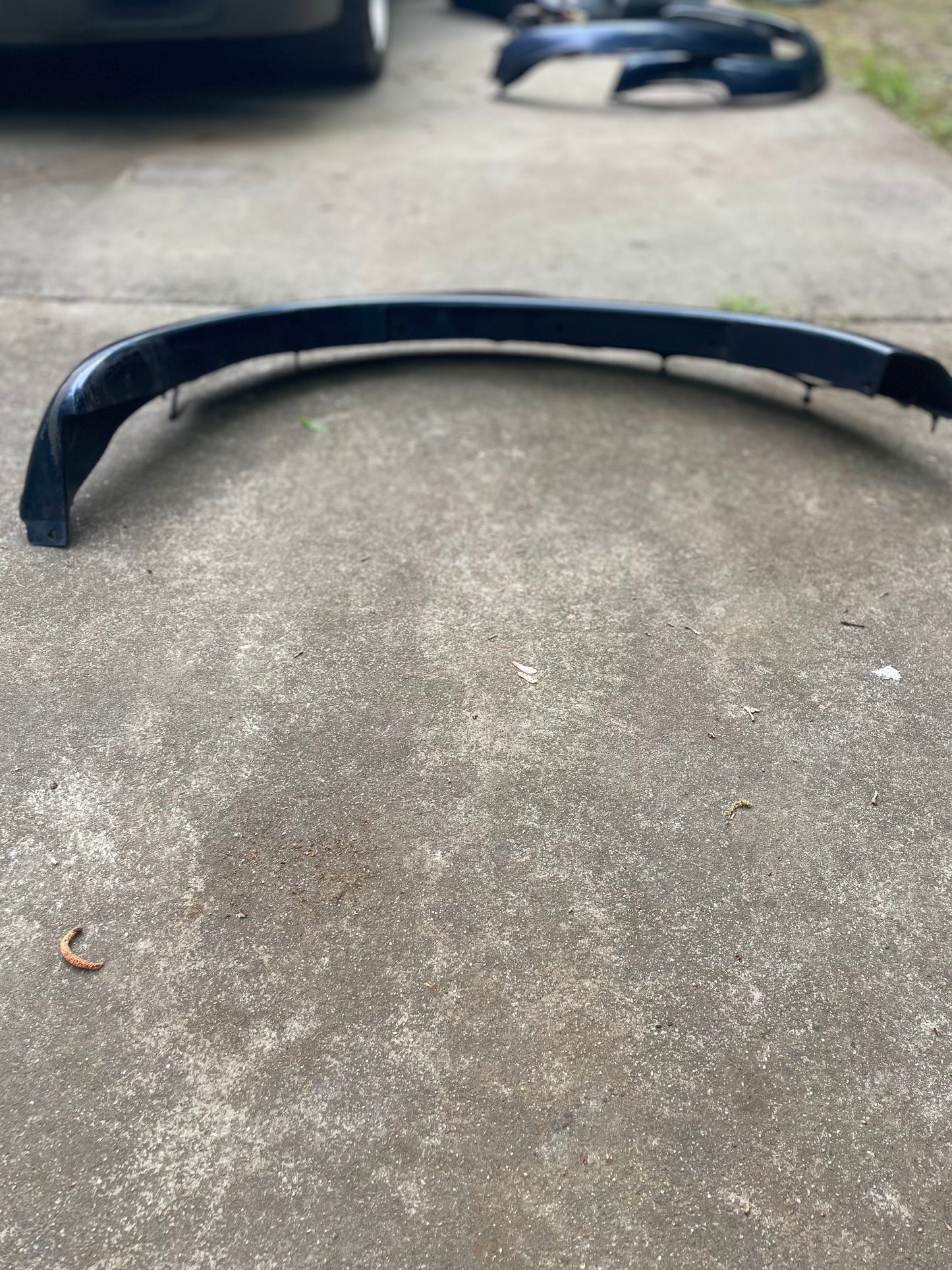 A black car bumper lies on a concrete driveway, with a General Motors OEM Front Right Passenger ZR2 Fender Flare Extension in Blue for 1994-2005 Chevy S10/GMC Sonoma visible in the blurred background.