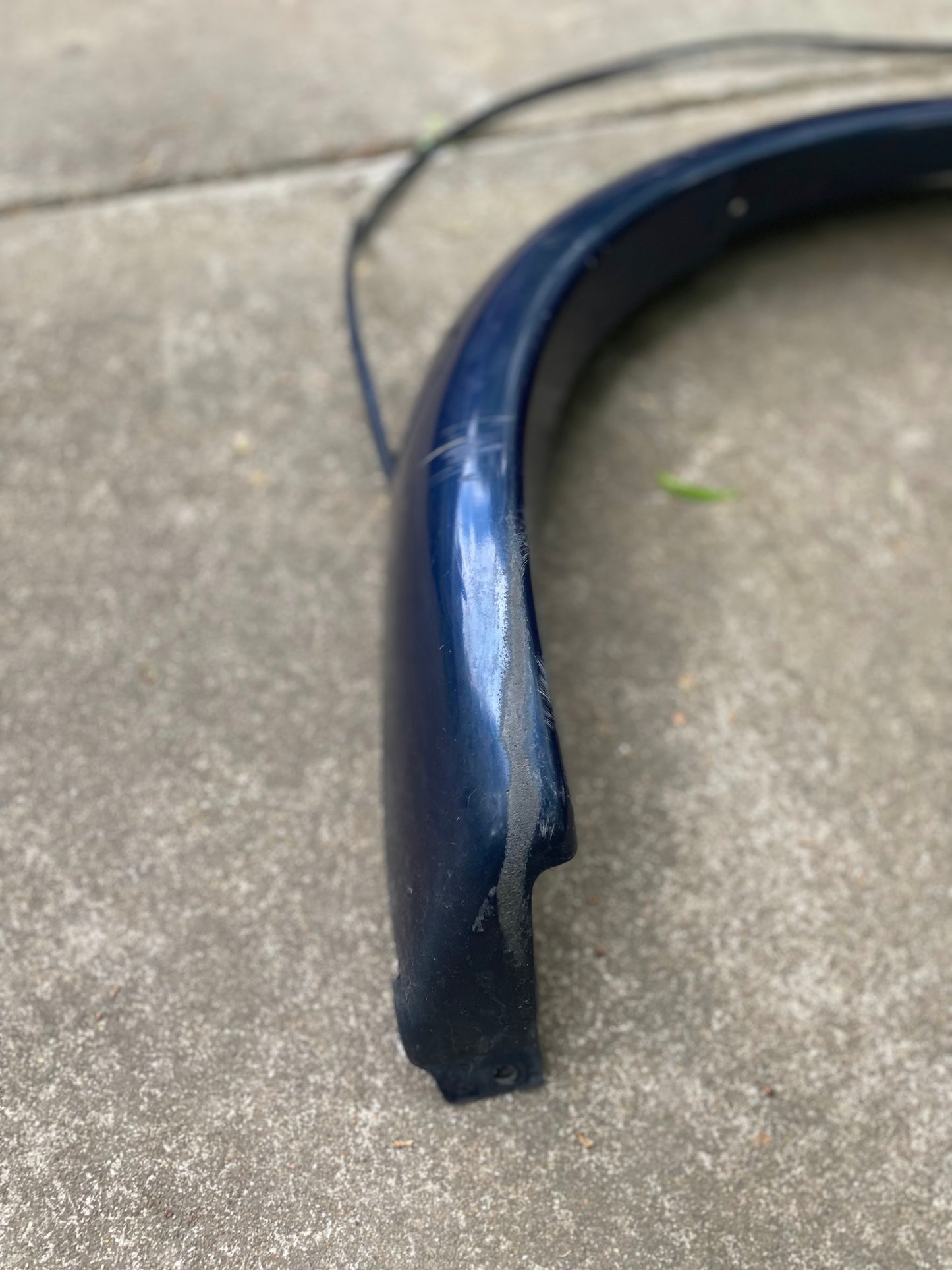 A close-up of a General Motors OEM Front Right Passenger ZR2 Fender Flare Extension in Blue for 1994-2005 Chevy S10 and GMC Sonoma, showing scratches and a crack, rests on concrete with a thin black cable and small green leaf nearby.
