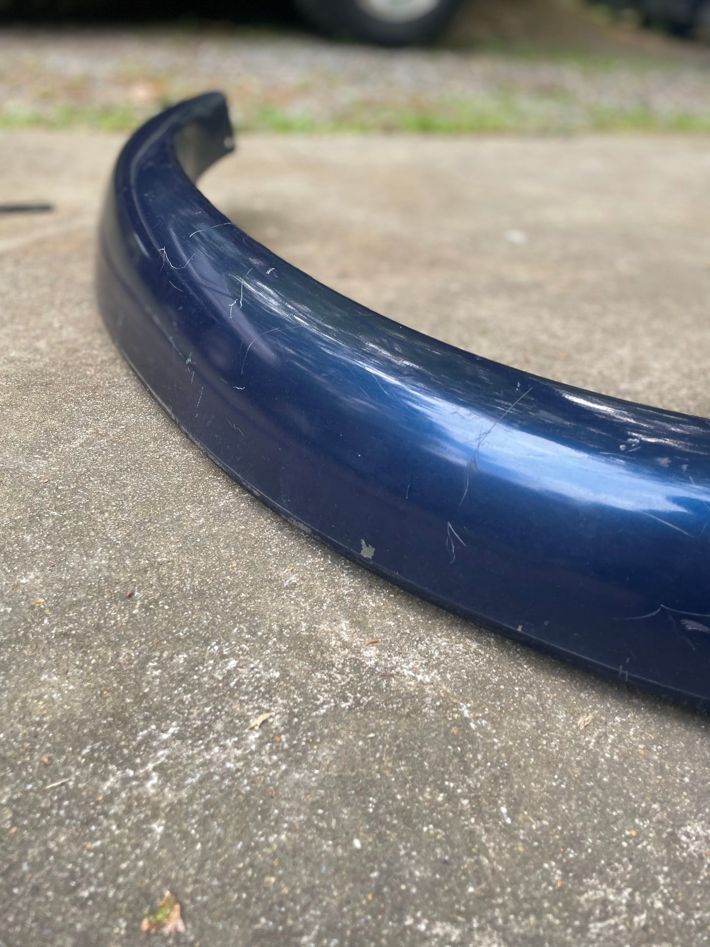 A close-up of a General Motors OEM Front Right Passenger ZR2 Fender Flare Extension in blue for 1994-2005 Chevy S10 and GMC Sonoma, showing visible scratches on a concrete surface with blurred grass and a vehicle tire in the background.