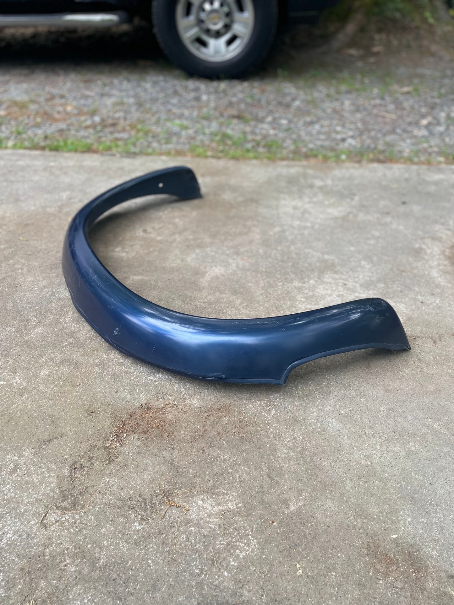 A blue OEM General Motors rear ZR2 bed flares extension pair for 1994-2004 Chevy S10 and GMC Sonoma is placed on a concrete surface outdoors, with a Chevrolet S10 ZR2 and gravel visible in the background.