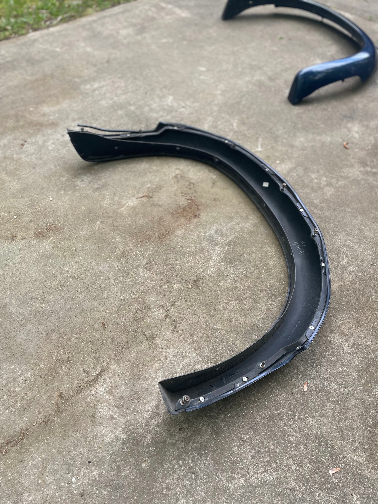 A curved black plastic car bumper lies on a concrete surface outdoors, with an OEM Pair Rear ZR2 Bed Flares Extension in Blue by General Motors for 1994-2004 Chevy S10 or GMC Sonoma visible in the background.