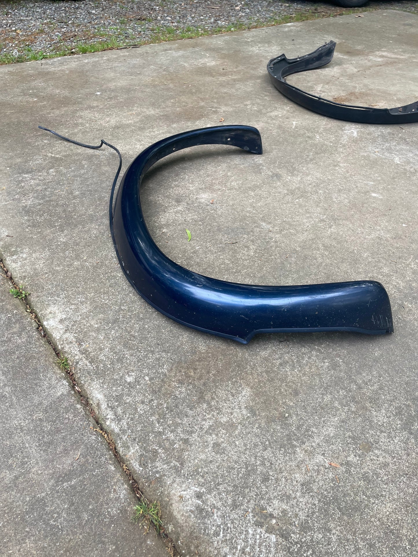 A blue OEM Pair Rear ZR2 Bed Flares Extension by General Motors for 1994-2004 Chevy S10 or GMC Sonoma lies outdoors on concrete, with another flare in the background. Grass and gravel are visible at the edges.
