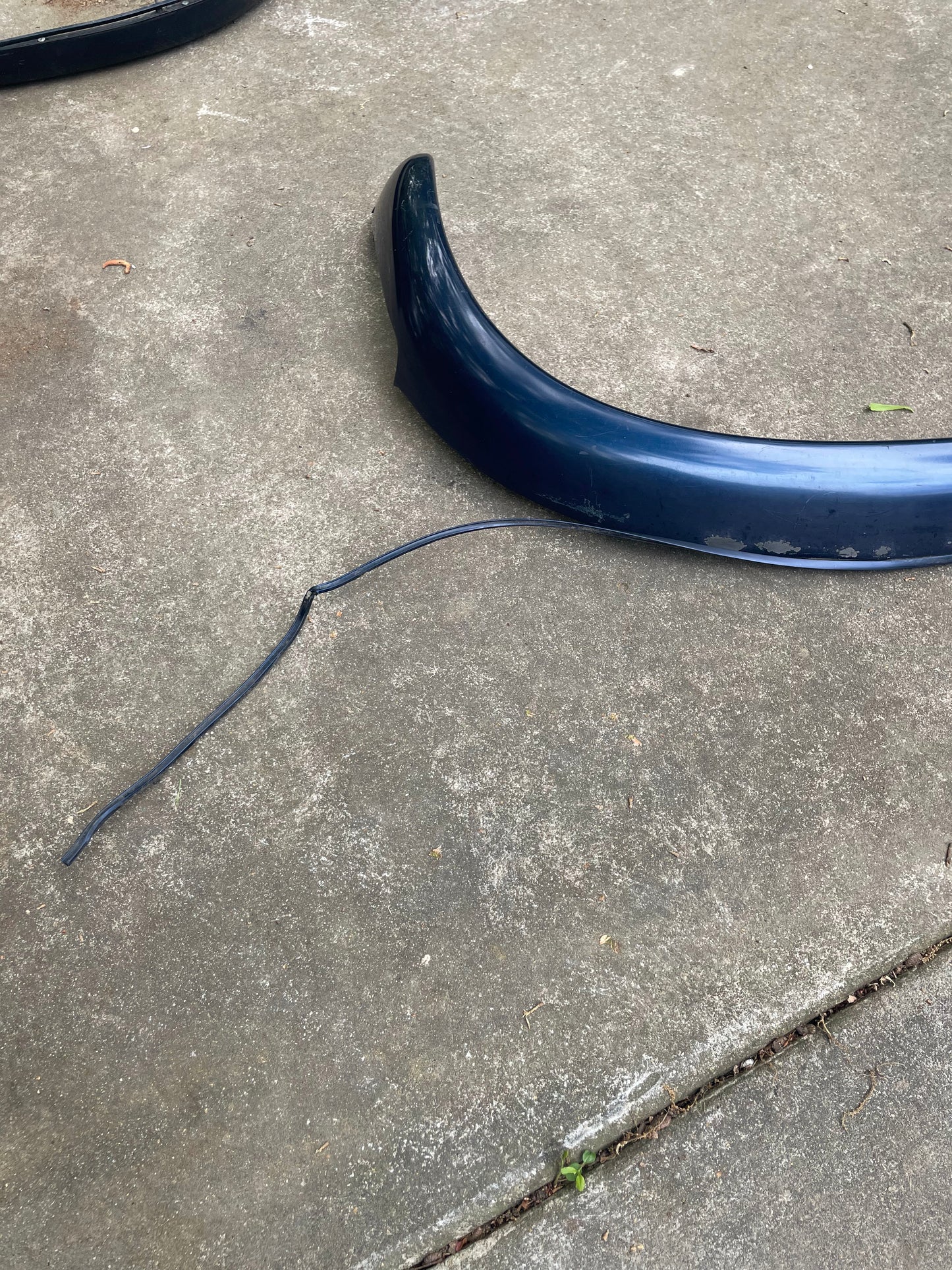 A pair of OEM General Motors Rear ZR2 Bed Flares Extension in blue for 1994-2004 Chevy S10 and GMC Sonoma, along with a thin black cable, are placed on a concrete driveway.