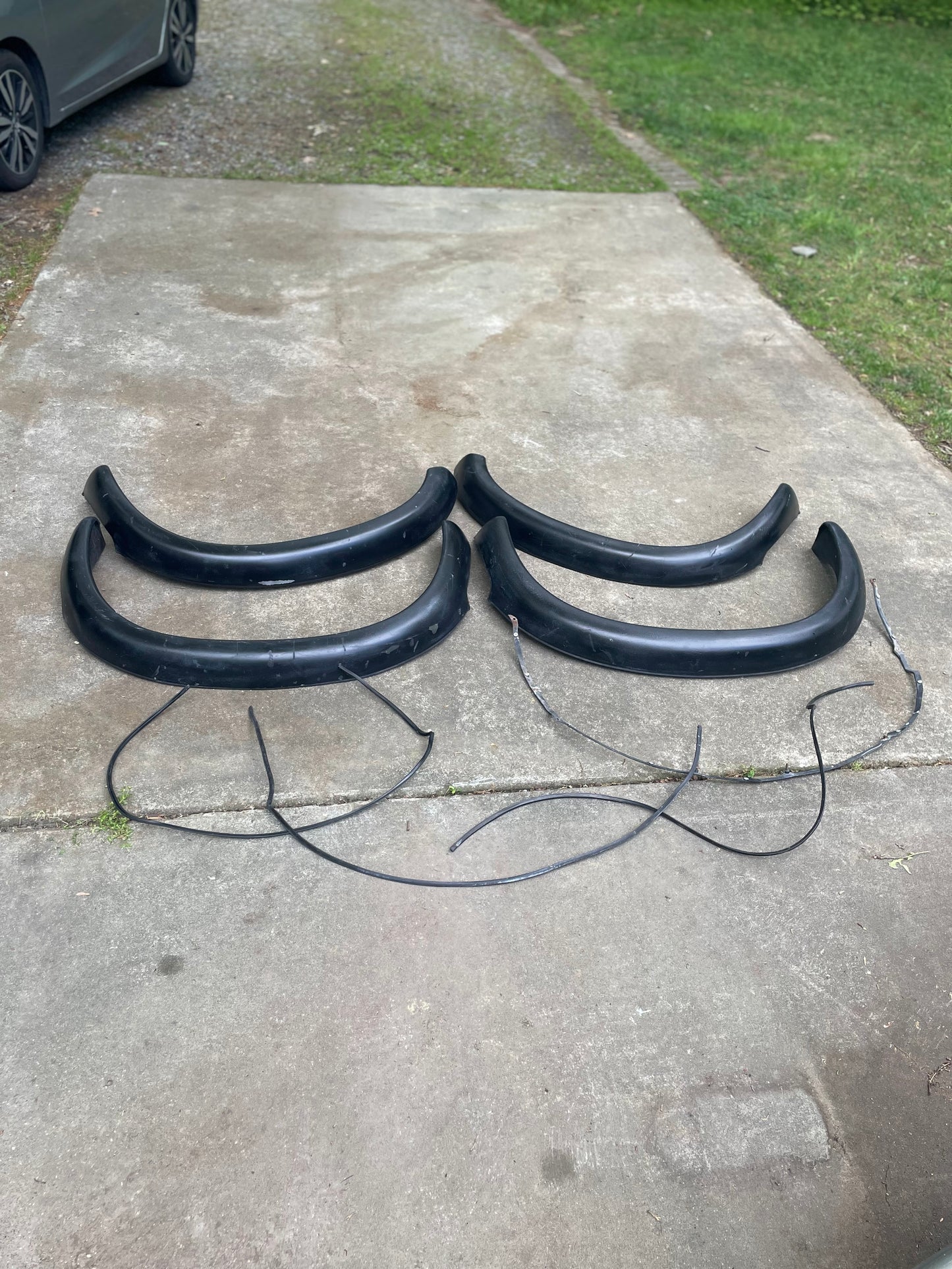A set of four black OEM ZR2 Fender Flares Extensions by General Motors for 1994-2004 Chevy S10 and GMC Sonoma are displayed on a concrete driveway, with grass and part of a silver Chevrolet S10 ZR2 visible.