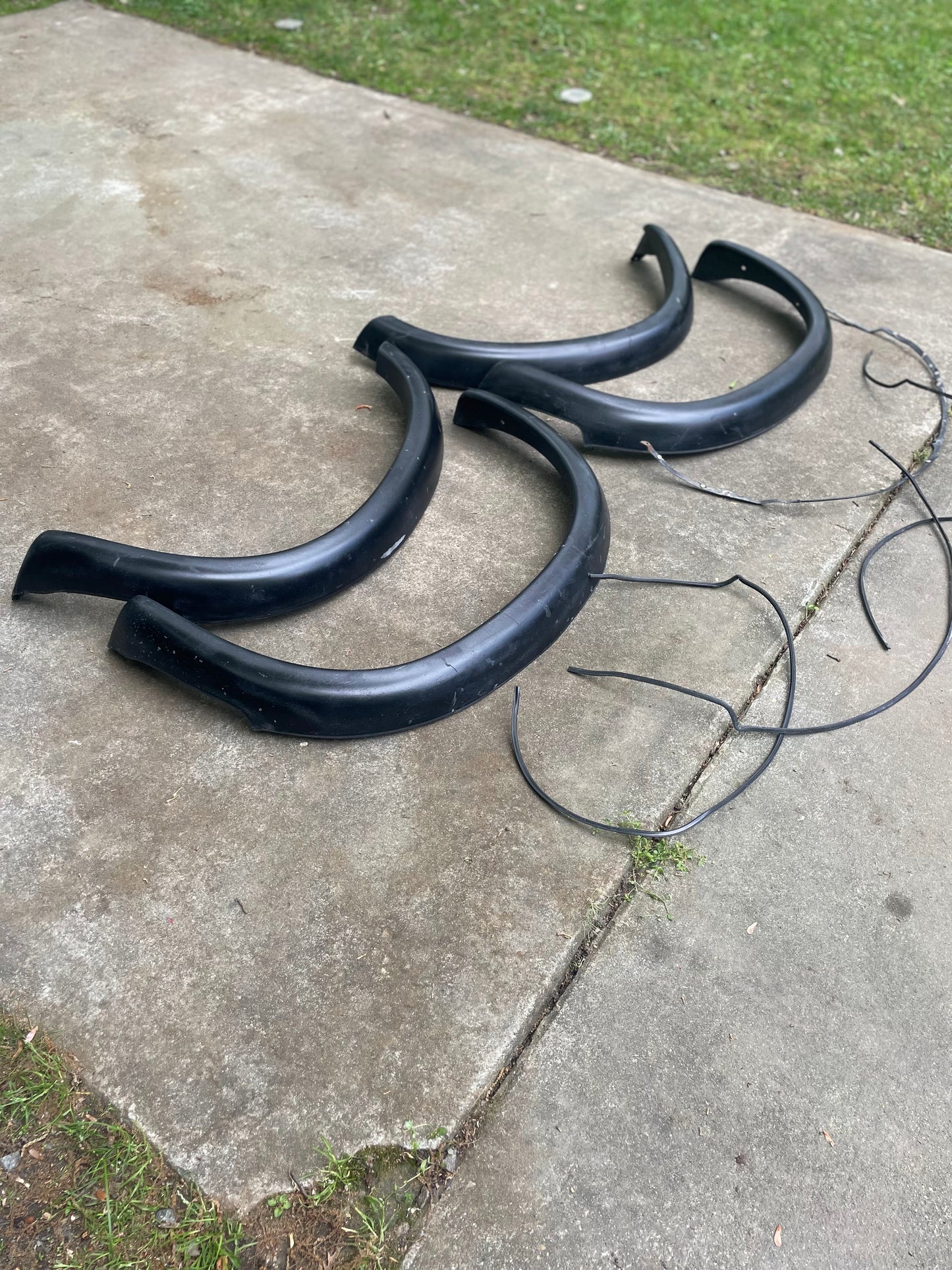 Three black plastic General Motors OEM ZR2 Fender Flares Extensions for 1994-2004 Chevy S10 or GMC Sonoma are shown with long rubber strips on a concrete surface outdoors, grass in the background.