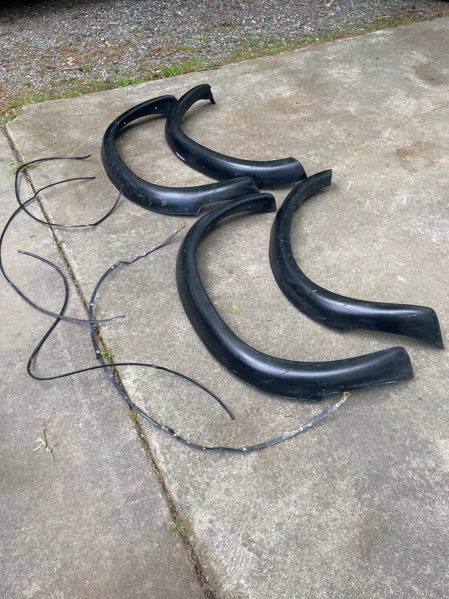 A General Motors OEM Set ZR2 Fender Flares Extension for 1994-2004 Chevy S10 or GMC Sonoma, including four black flares and two trim strips, is laid out on a concrete driveway with gravel and grass in the background.
