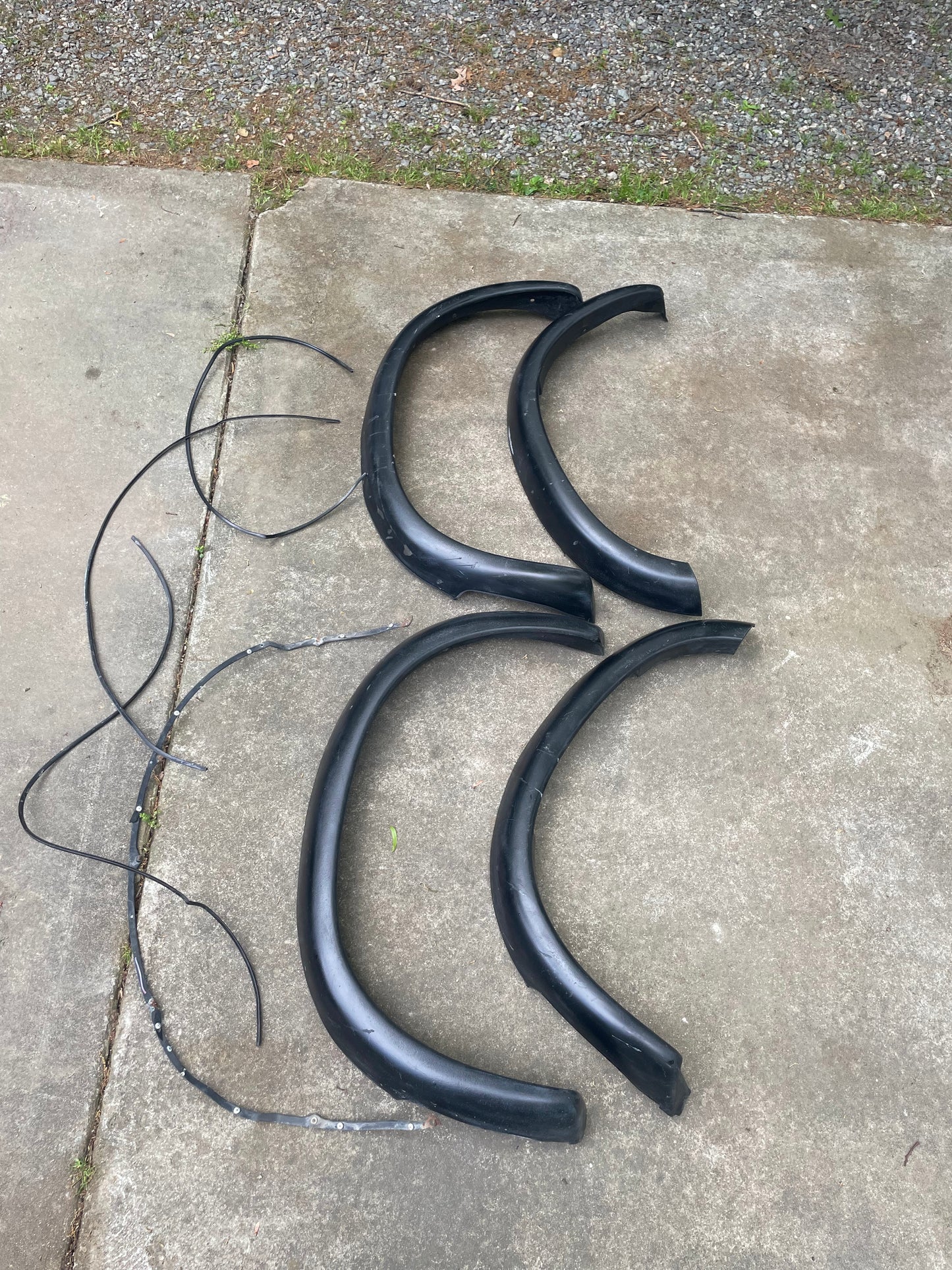 Four black General Motors OEM Set ZR2 Fender Flares Extensions for 1994-2004 Chevy S10 or GMC Sonoma, plus two rubber trim strips, laid out on a concrete driveway with gravel and grass visible at the top.