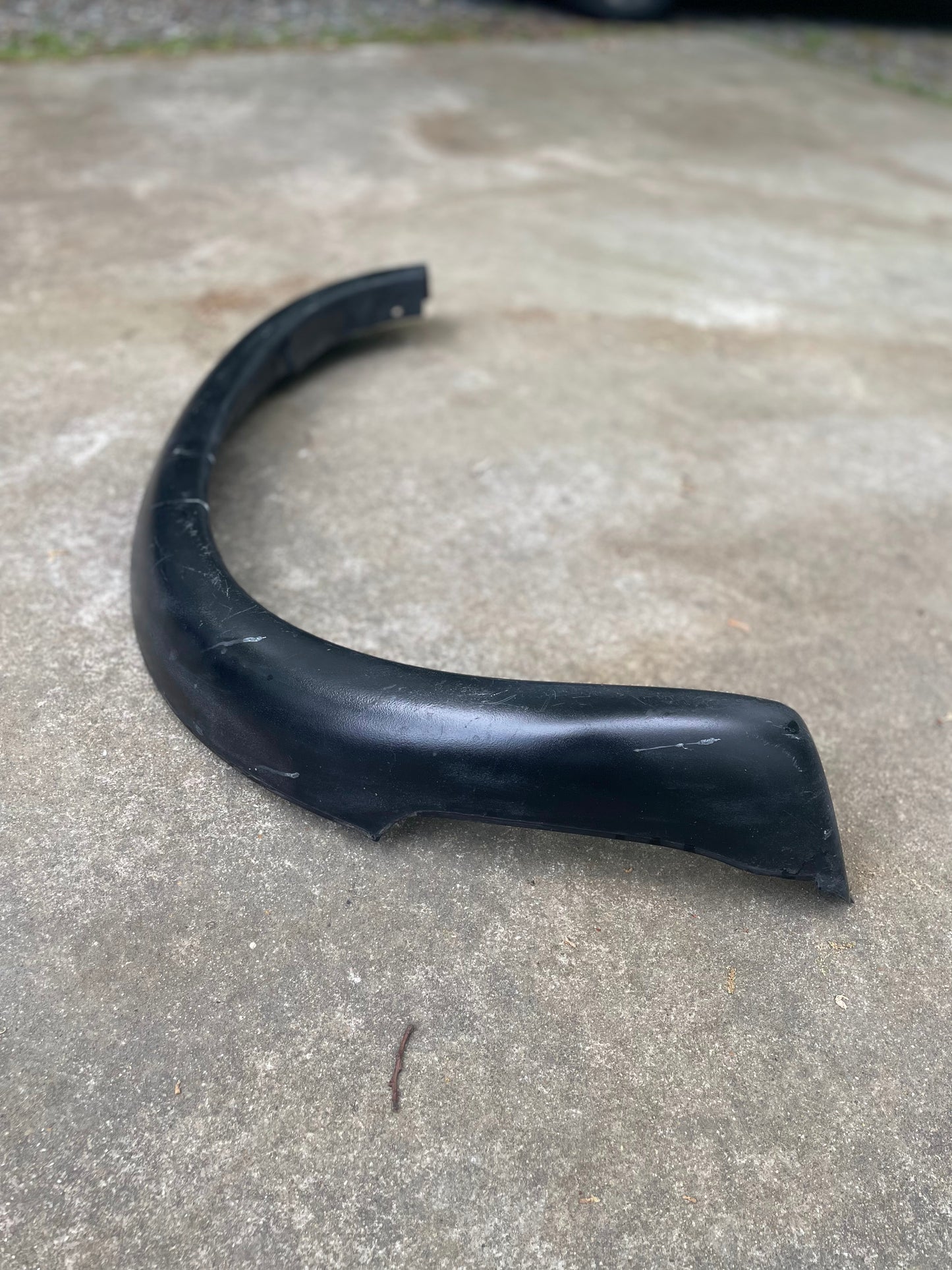 A General Motors OEM ZR2 Fender Flare Extension for 1994-2004 Chevy S10 or GMC Sonoma, black and curved, is lying on a concrete surface outdoors.
