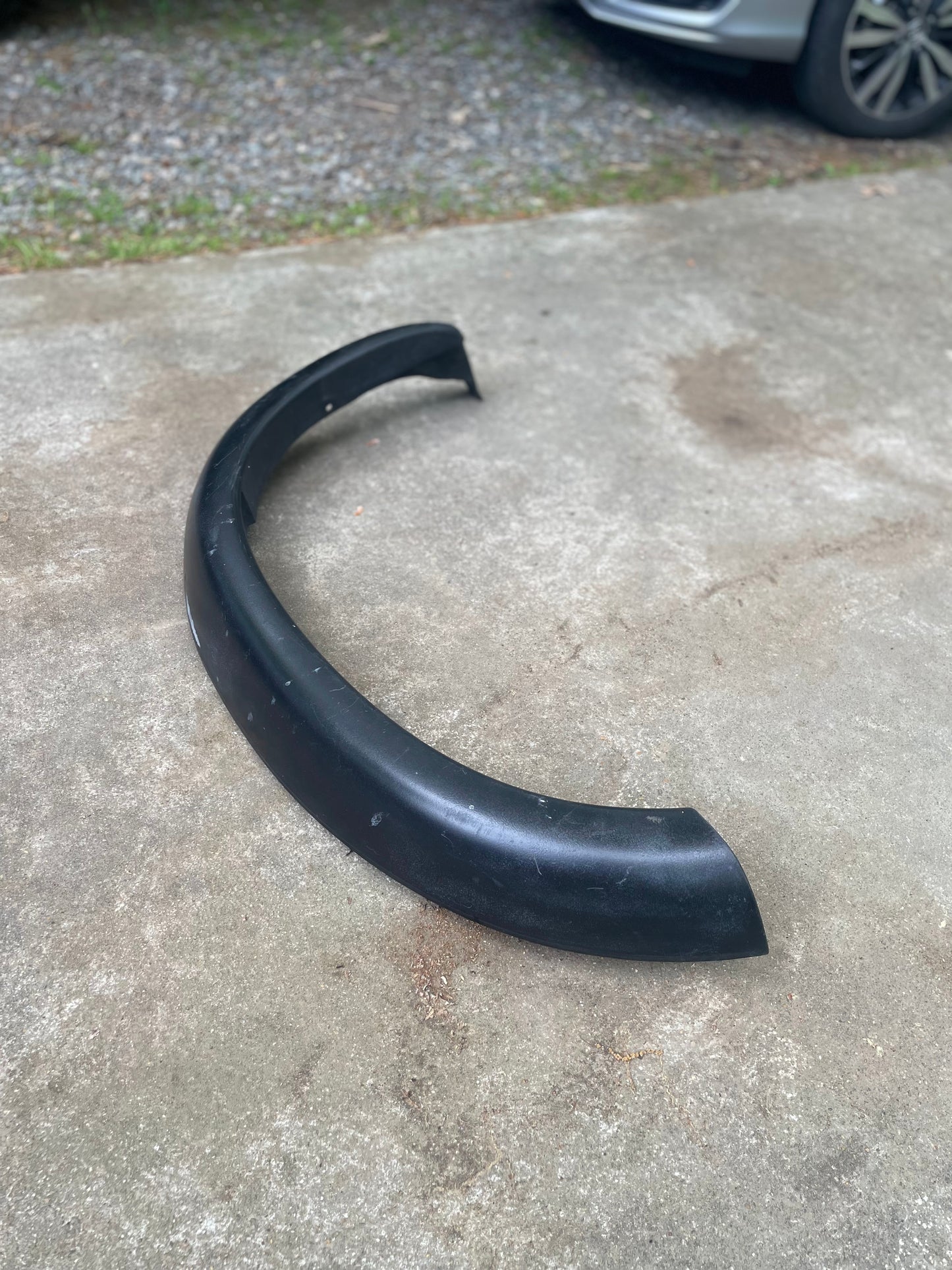 A black car bumper lies on a concrete driveway near a General Motors OEM Set ZR2 Fender Flares Extension for 1994-2004 Chevy S10, GMC Sonoma, with cars—including a Chevrolet S10 ZR2—and gravel visible in the background.