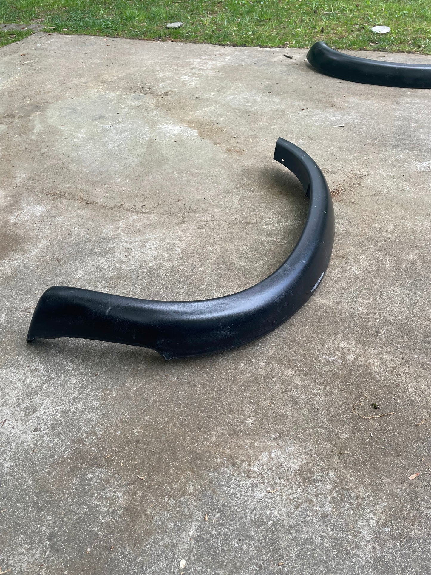 A black General Motors OEM Set ZR2 Fender Flares Extension for 1994-2004 Chevy S10 and GMC Sonoma rests outdoors on concrete, with grass behind it. Another matching fender flare is partly visible in the top right corner.
