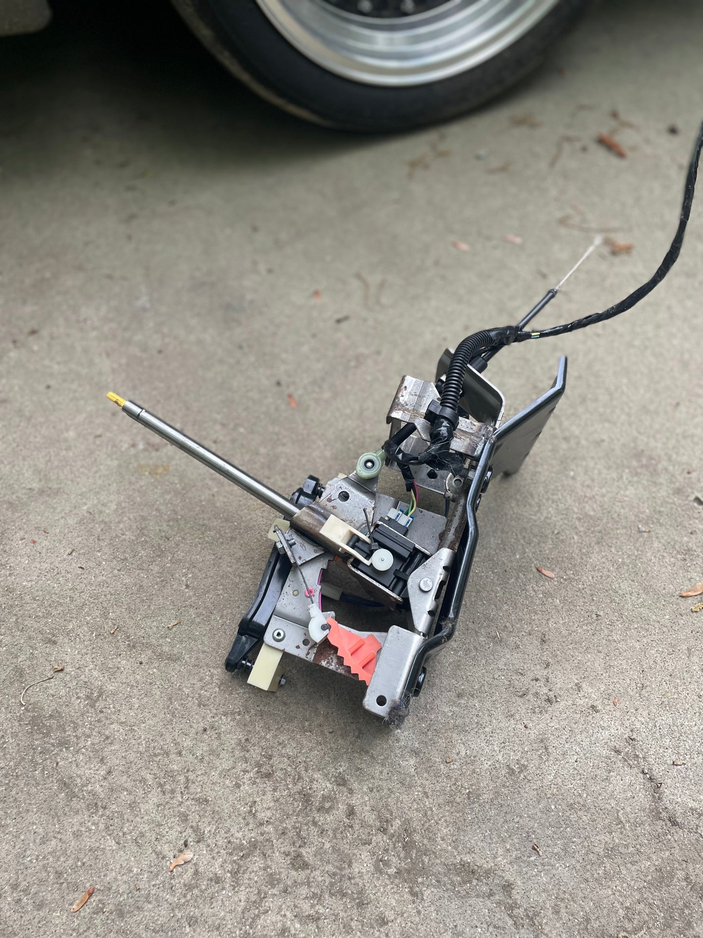 A close-up of a disassembled General Motors KIT Center Shift Console Swap with woodgrain in Graphite for 1994-2004 Chevy S10, GMC Sonoma, and more, showing exposed gears and wires on concrete near a car tire.