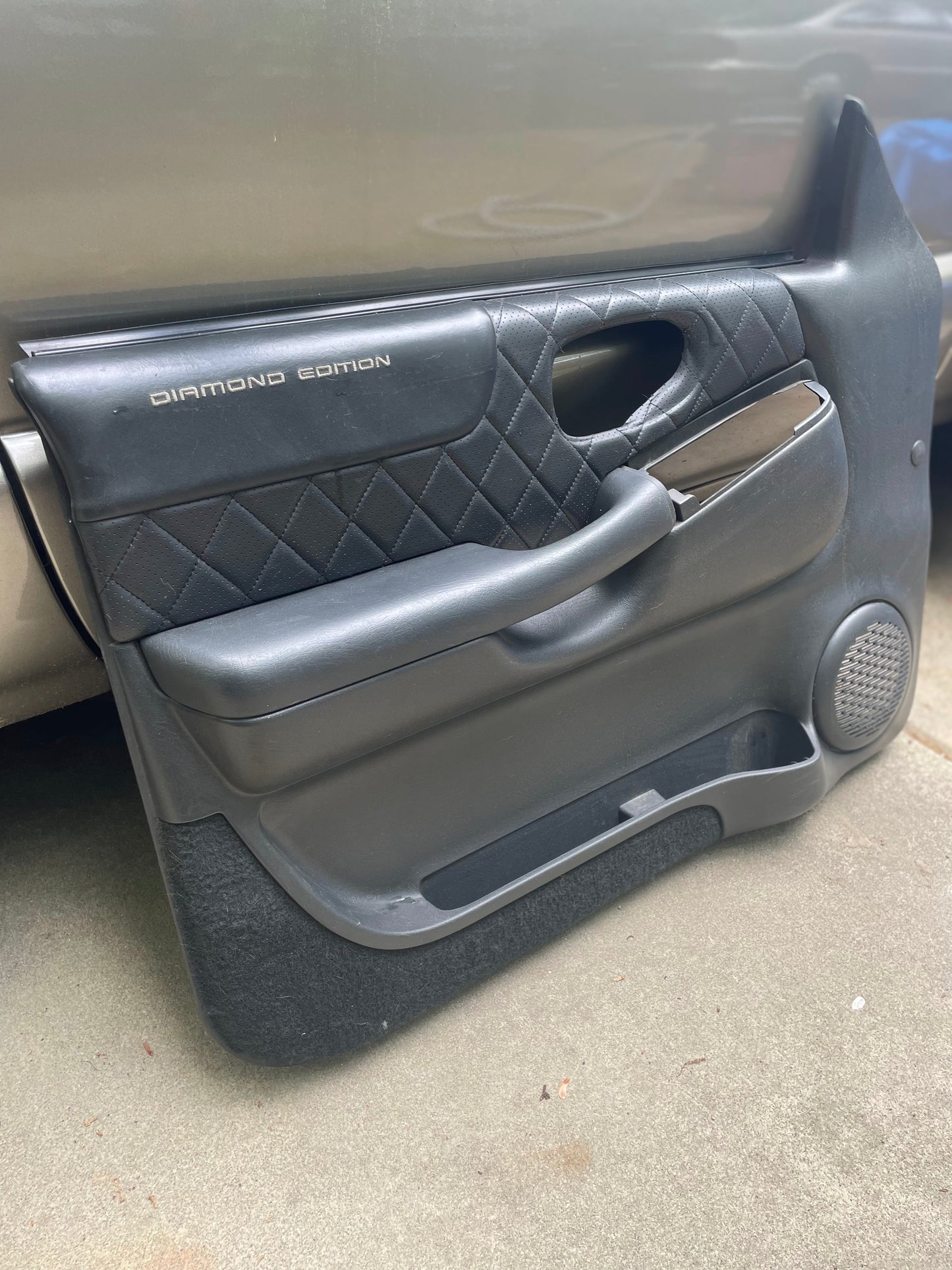 A pair of General Motors OEM front power leather diamond door panels in dark gray, designed for 1998-2005 Chevy S10 and GMC Sonoma, rests on a concrete surface near a silver vehicle.