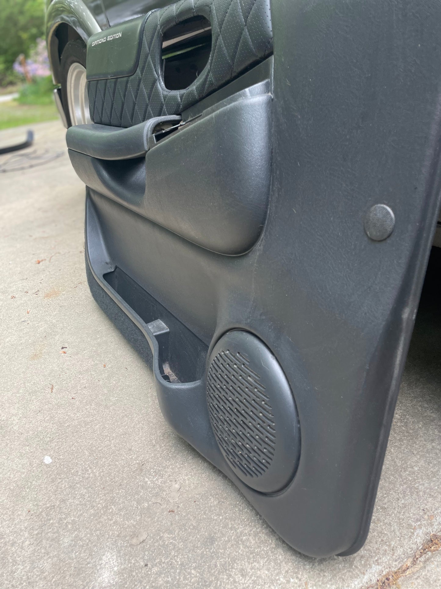 Close-up of an OEM pair of front power leather diamond door panels in dark gray from General Motors for 1998-2005 Chevy S10, GMC Sonoma, and more, showing speaker, storage compartment, quilted paneling, and part of a tire on concrete.