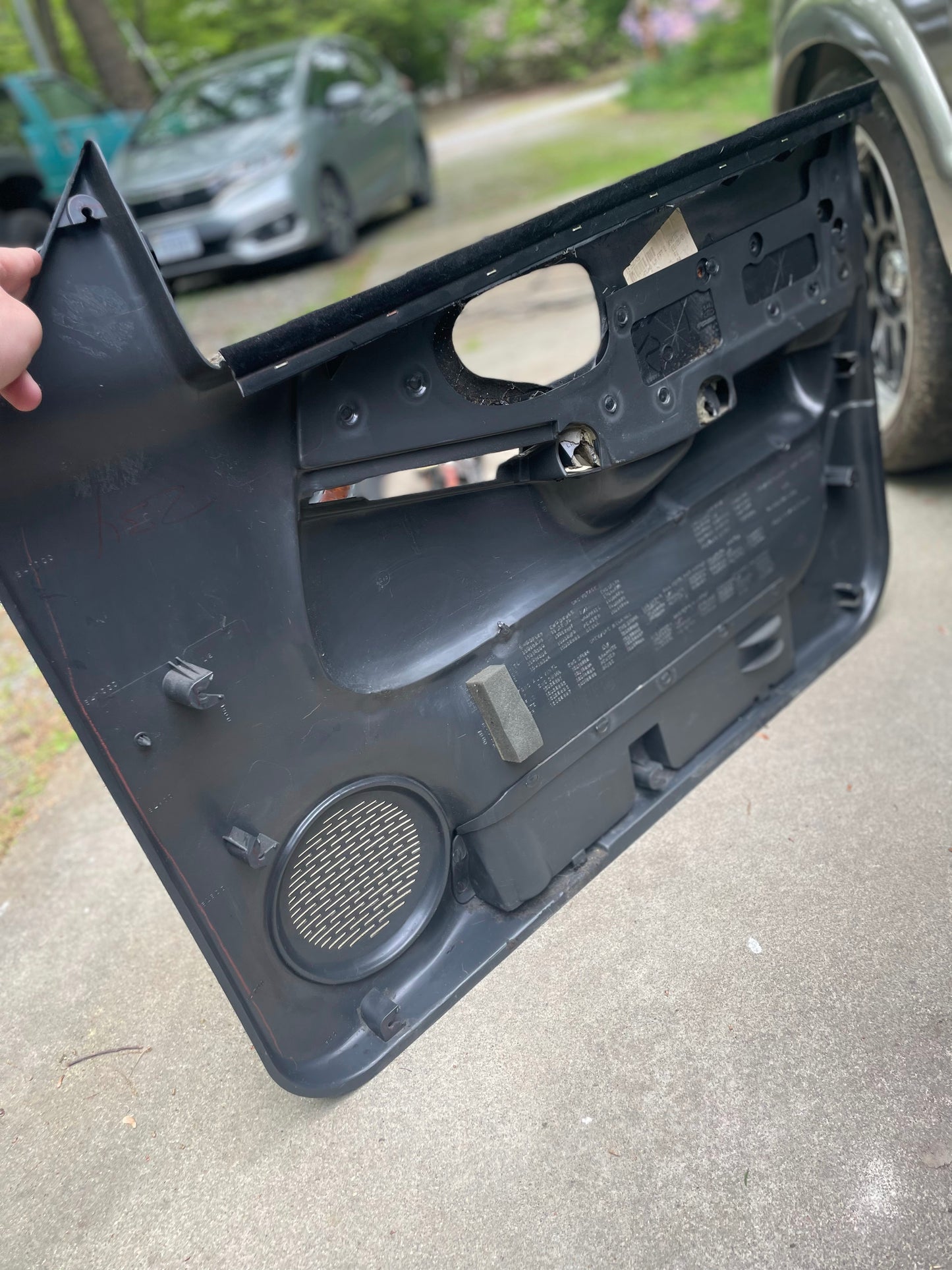 Someone holds a General Motors OEM pair of front power leather diamond door panels in dark gray for 1998-2005 Chevy S10 and GMC Sonoma on a concrete driveway, with cars and greenery blurred in the background.