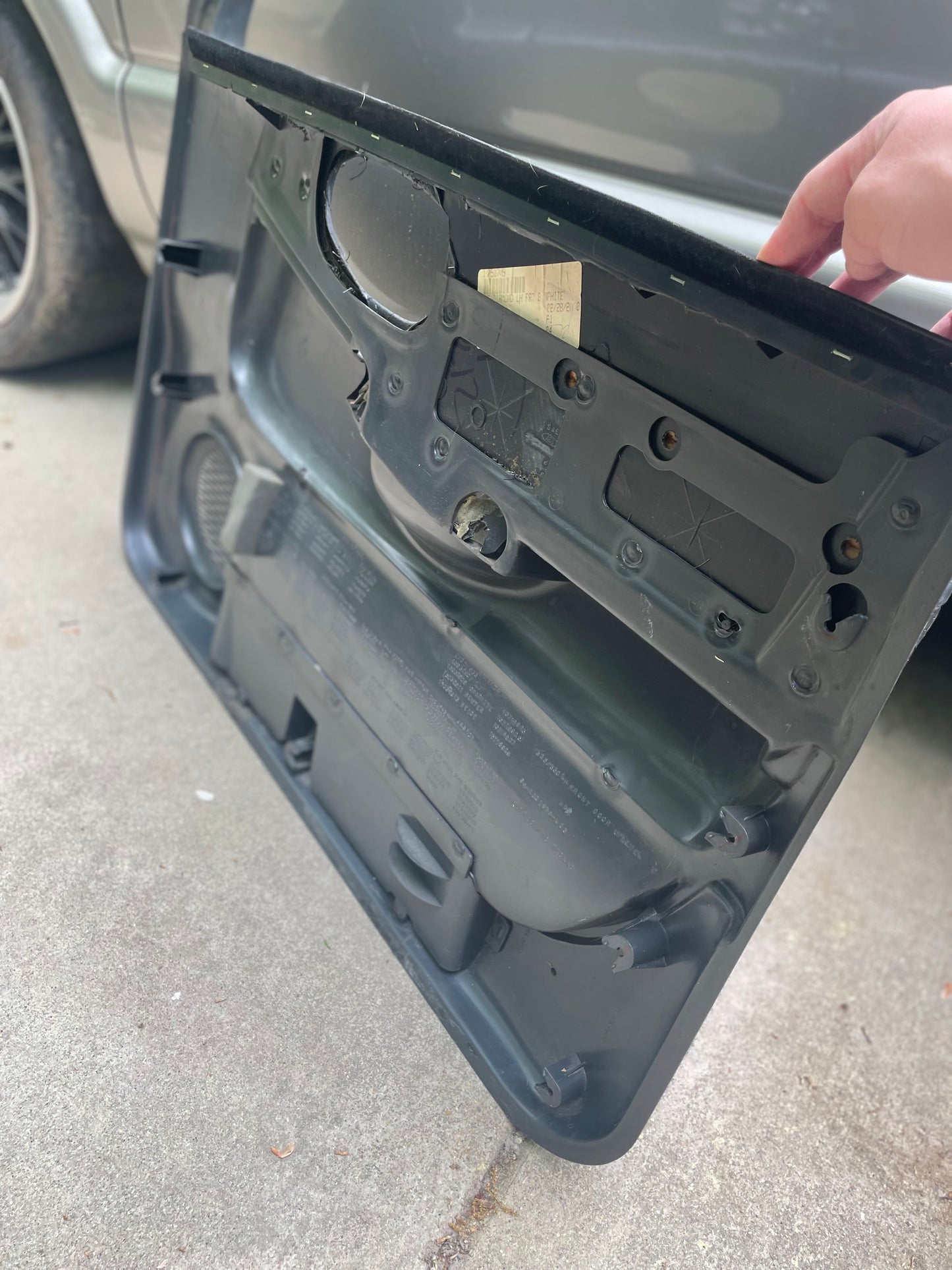 A person holds a detached General Motors OEM pair of front power leather diamond door panels in dark gray, designed for 1998-2005 Chevy S10 and GMC Sonoma, next to a silver car on a concrete driveway.