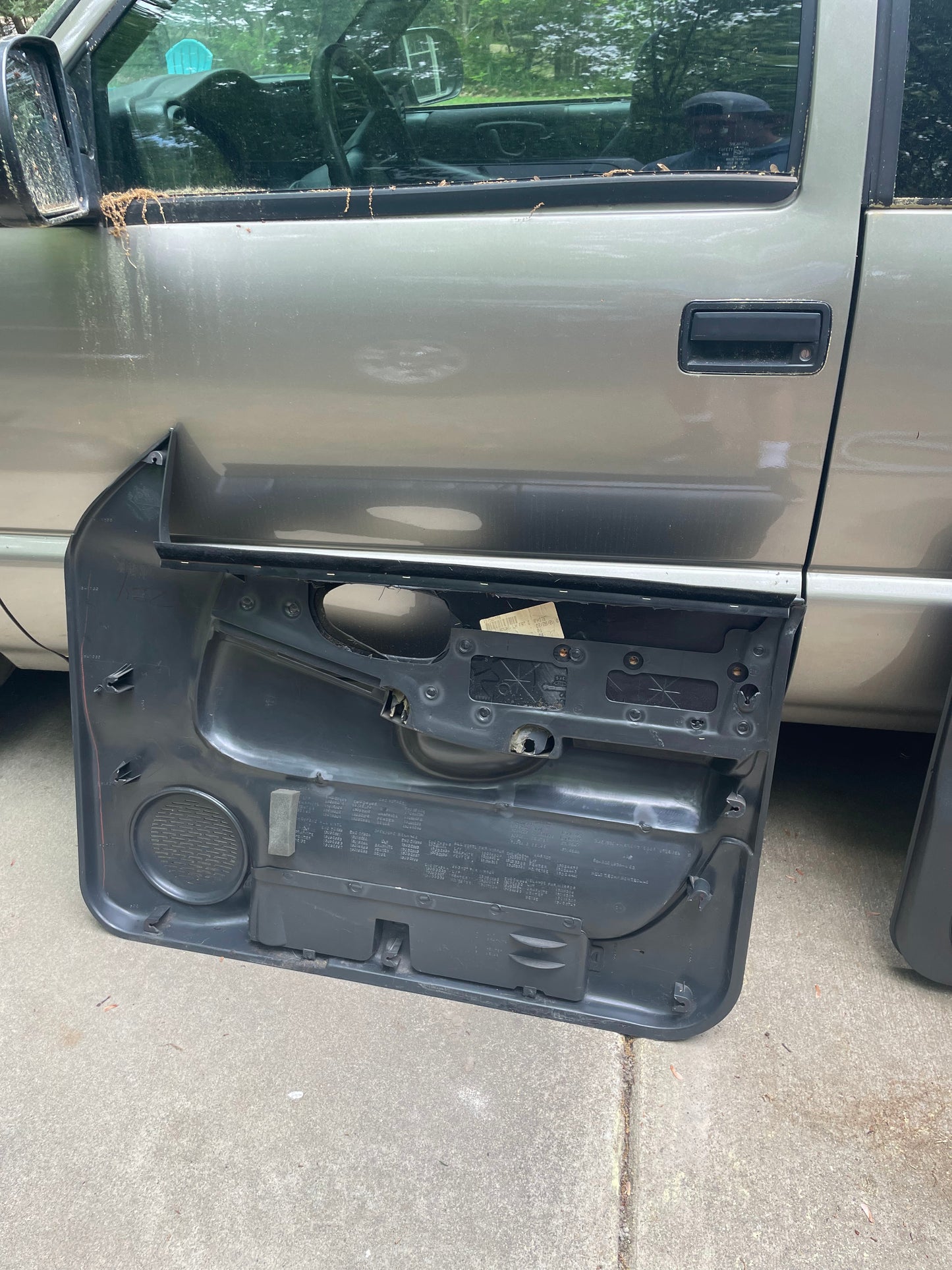 A General Motors pickup, possibly a 1998-2005 GMC Sonoma, is parked in a driveway. Its driver-side OEM Dark Gray Diamond Power Leather Door Panel is removed, exposing inner mechanisms and wiring; trees are reflected in the window.