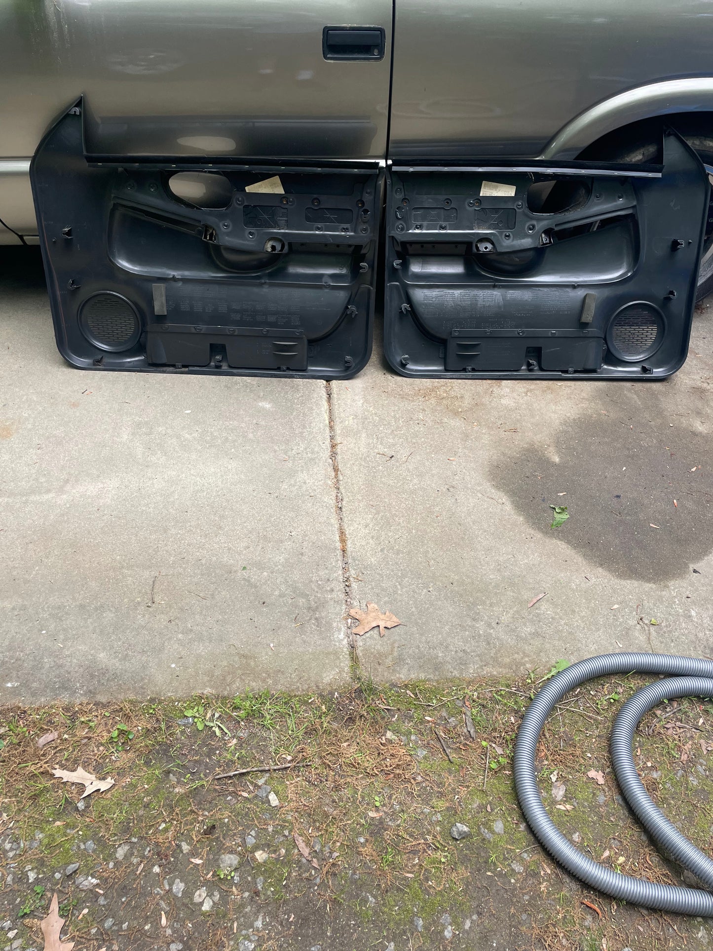 A pair of General Motors OEM front power leather diamond door panels in dark gray for 1998-2005 Chevy S10 and GMC Sonoma are side by side on a driveway before a silver vehicle, with a vacuum hose coiled to the right and leaves nearby.