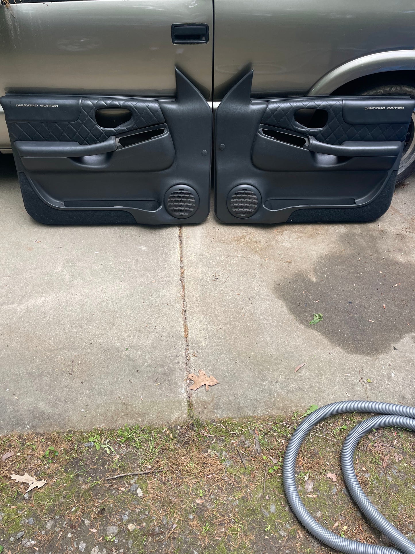 A pair of General Motors OEM dark gray leather diamond front power door panels for 1998-2005 Chevy S10/GMC Sonoma lean against a tan vehicle on a concrete driveway, with a vacuum hose seen in the lower right corner.