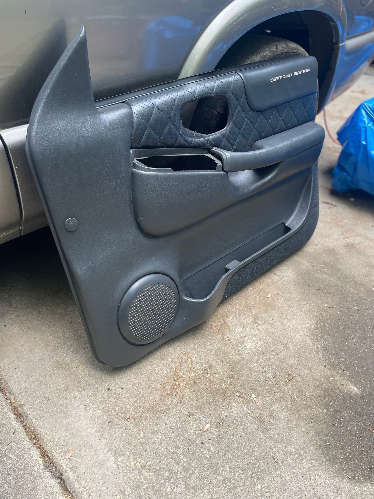 An OEM pair of General Motors front power leather diamond door panels in dark gray for 1998-2005 Chevy S10 and GMC Sonoma, shown upright on concrete beside a silver vehicle with a blue tarp visible in the background.