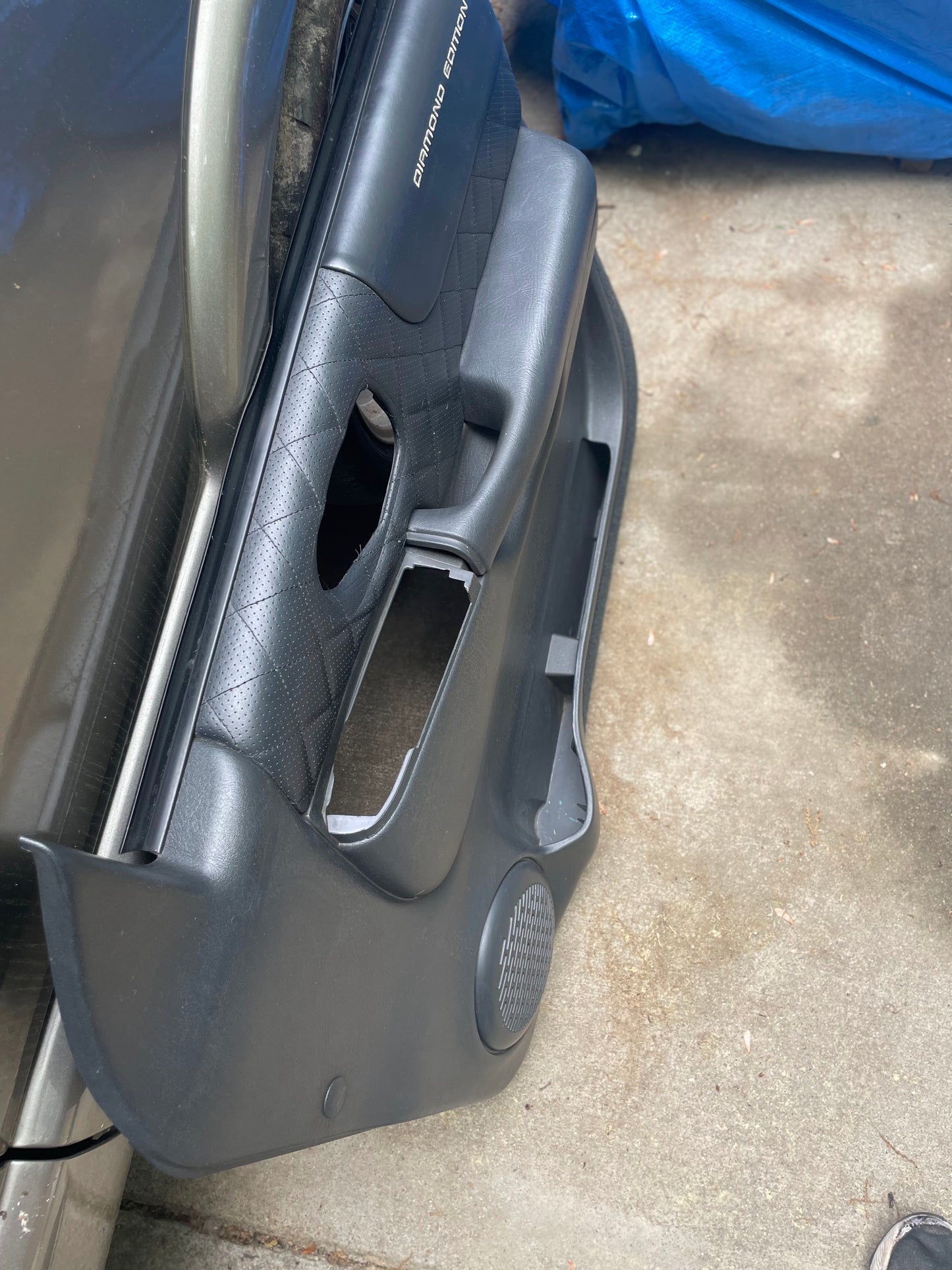 A General Motors OEM pair of front power leather diamond door panels in dark gray for 1998-2005 Chevy S10, GMC Sonoma, and more is shown on concrete with a missing handle section and a blue tarp in the background.