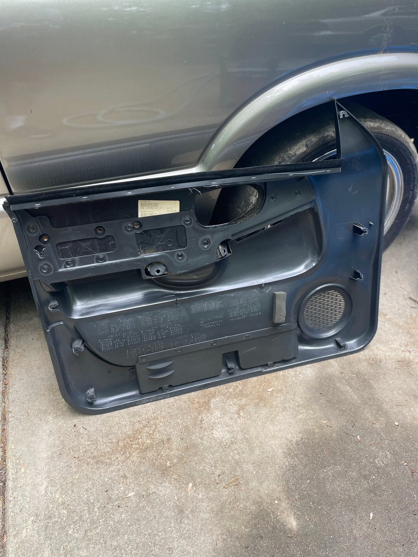 A General Motors OEM pair of front power leather diamond door panels in dark gray for 1998-2005 Chevy S10 and GMC Sonoma rests against the rear tire and fender of a silver vehicle on a concrete driveway, showing inner mechanisms and speaker.