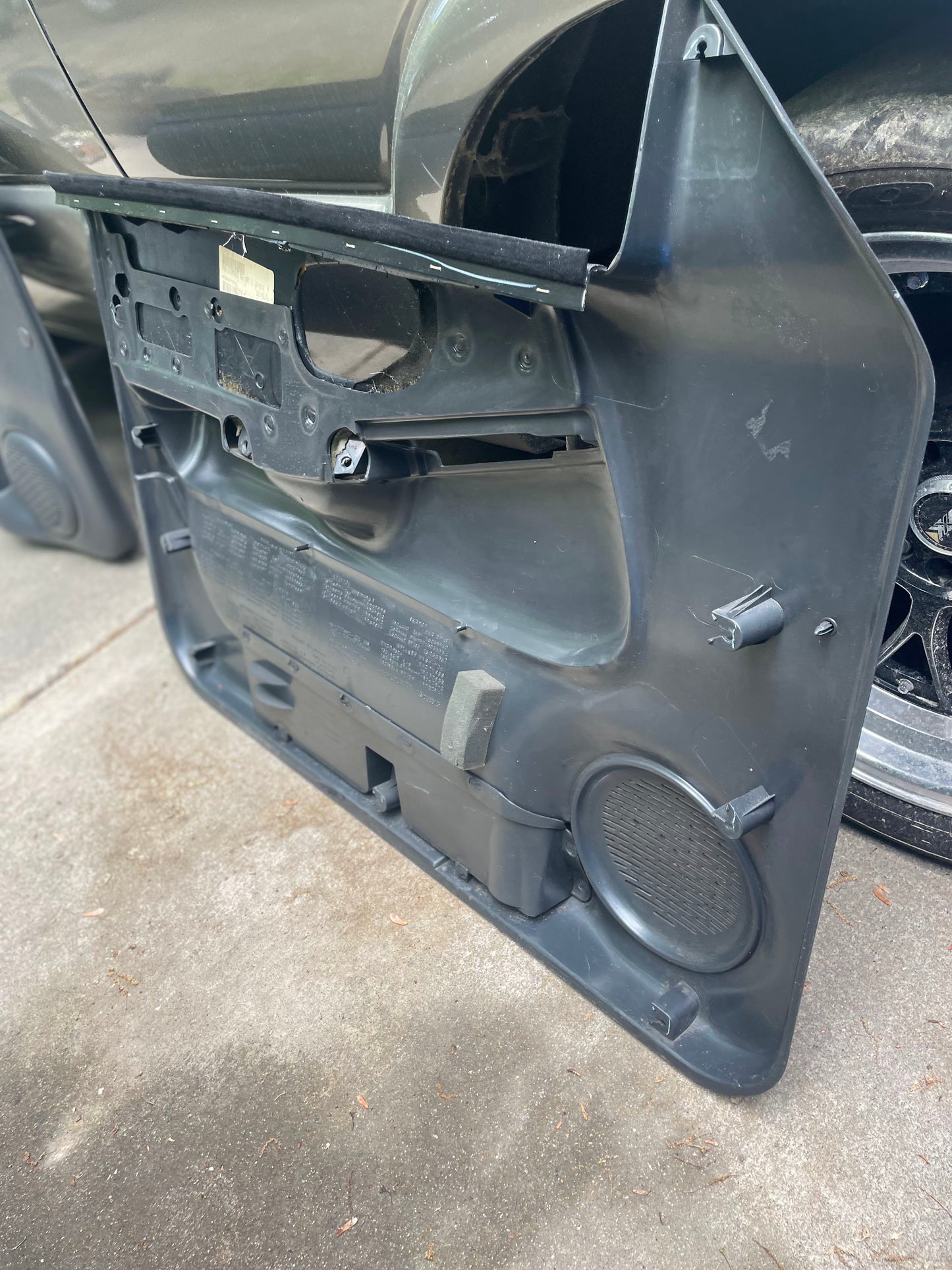 A pair of General Motors OEM front power leather diamond door panels in dark gray for 1998-2005 Chevy S10 and GMC Sonoma rests on the ground by a car, showing the interior side with mounting points and speaker housing.