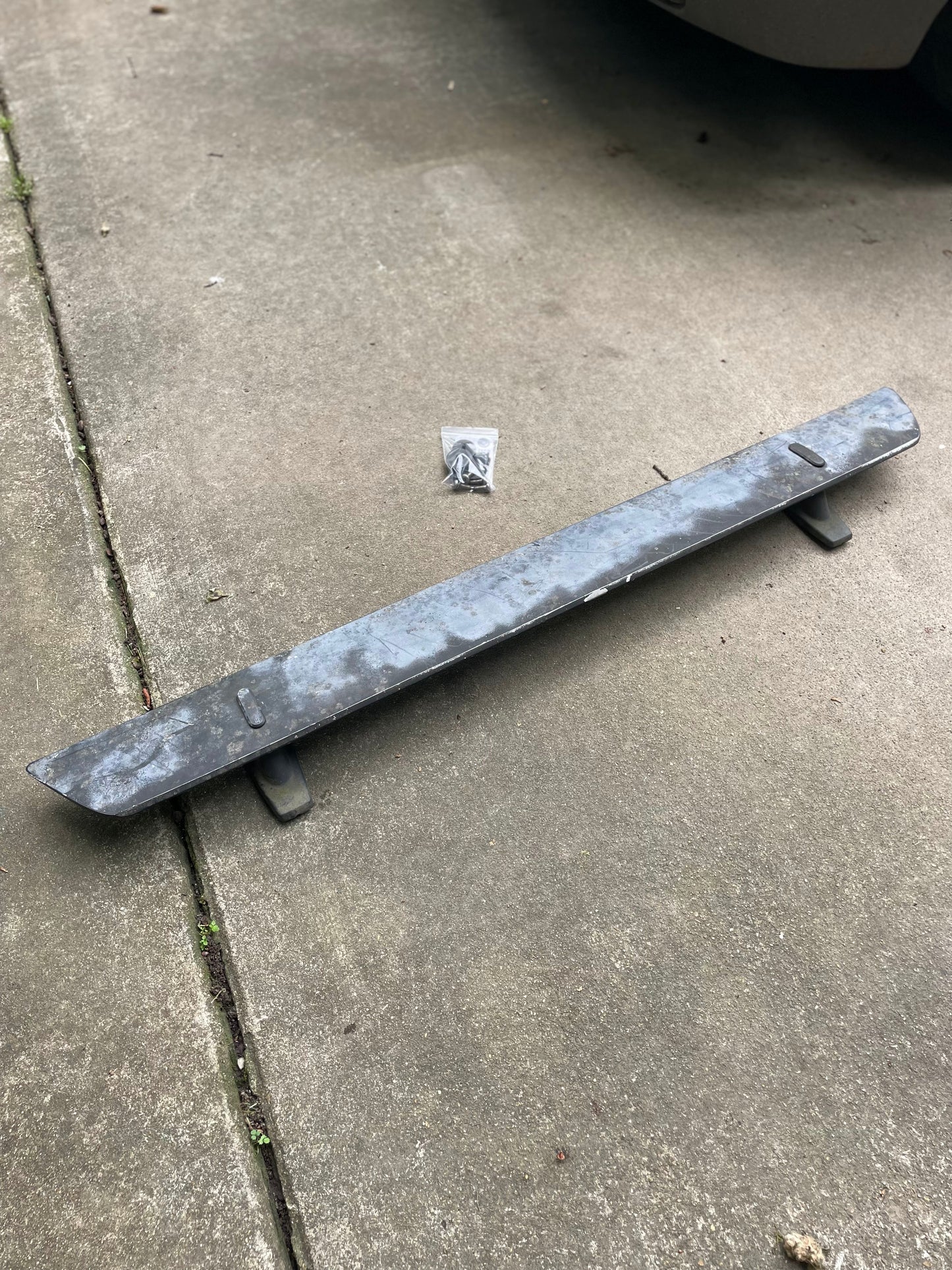 A General Motors OEM Rear Hatch Hinges with Spoiler Wing for 1983-1994 Chevrolet S10 Blazer sits on a concrete driveway, bolts in a small bag above it. Part of a vehicle is visible at the top right.