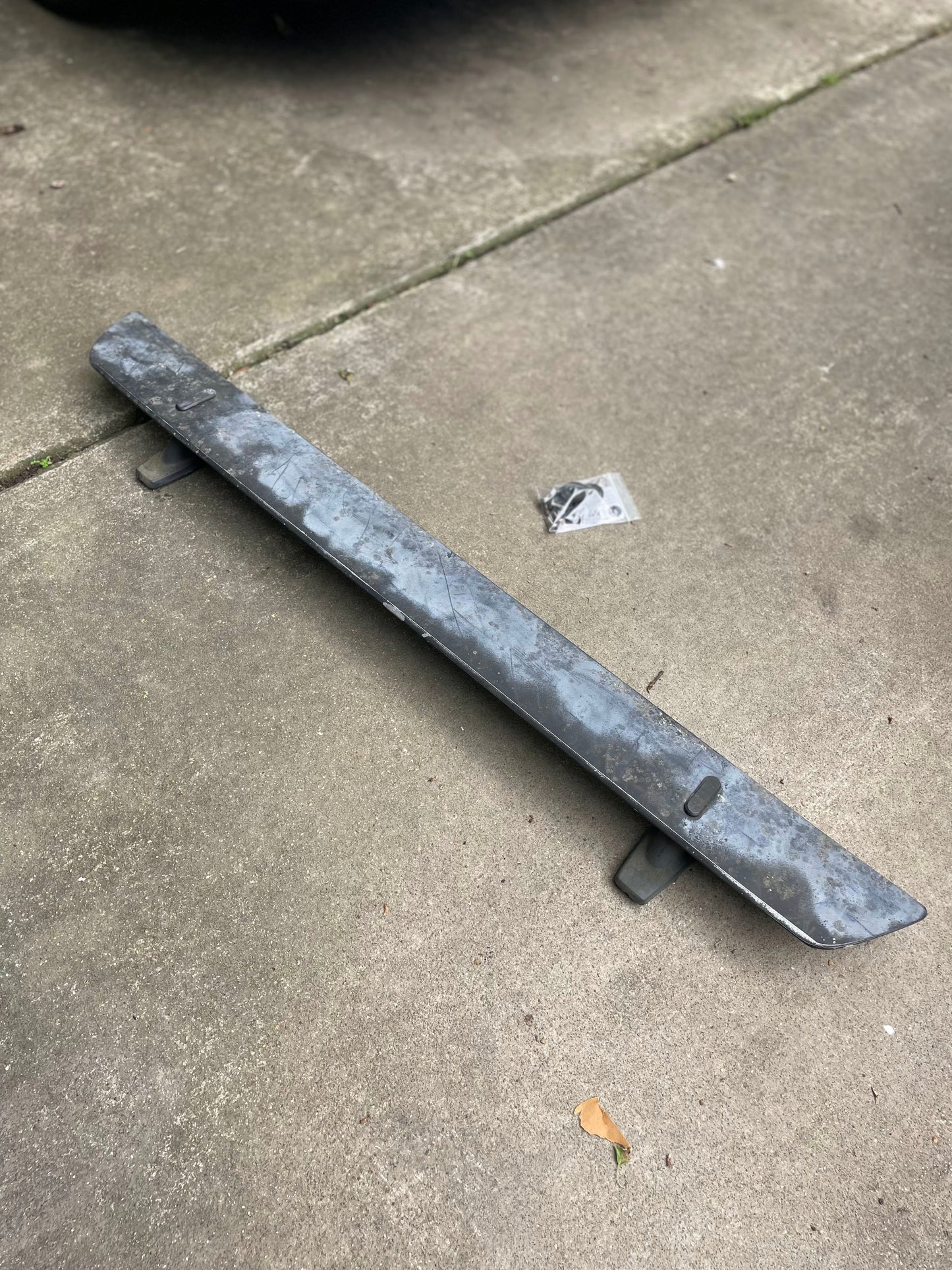General Motors OEM Rear Hatch Hinges with Spoiler Wing for 1983-1994 Chevrolet S10 Blazer rest on a concrete driveway, accompanied by a small bag of screws; scattered leaves are also visible nearby.